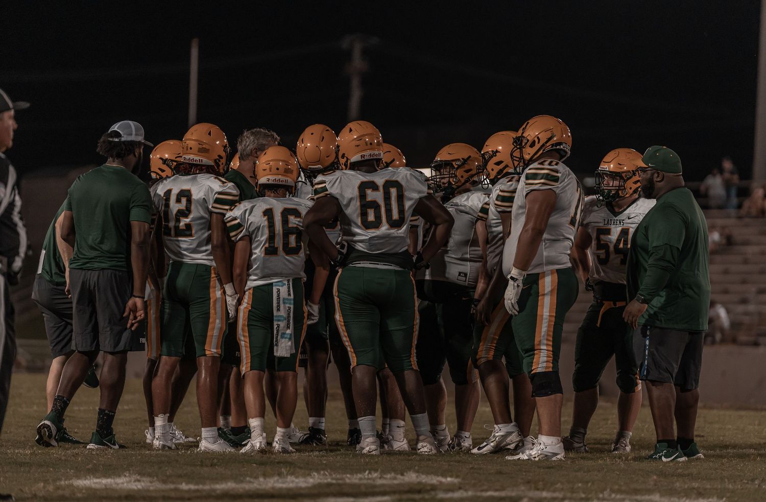 High school football team huddles together on field in green and orange uniforms during night game.
