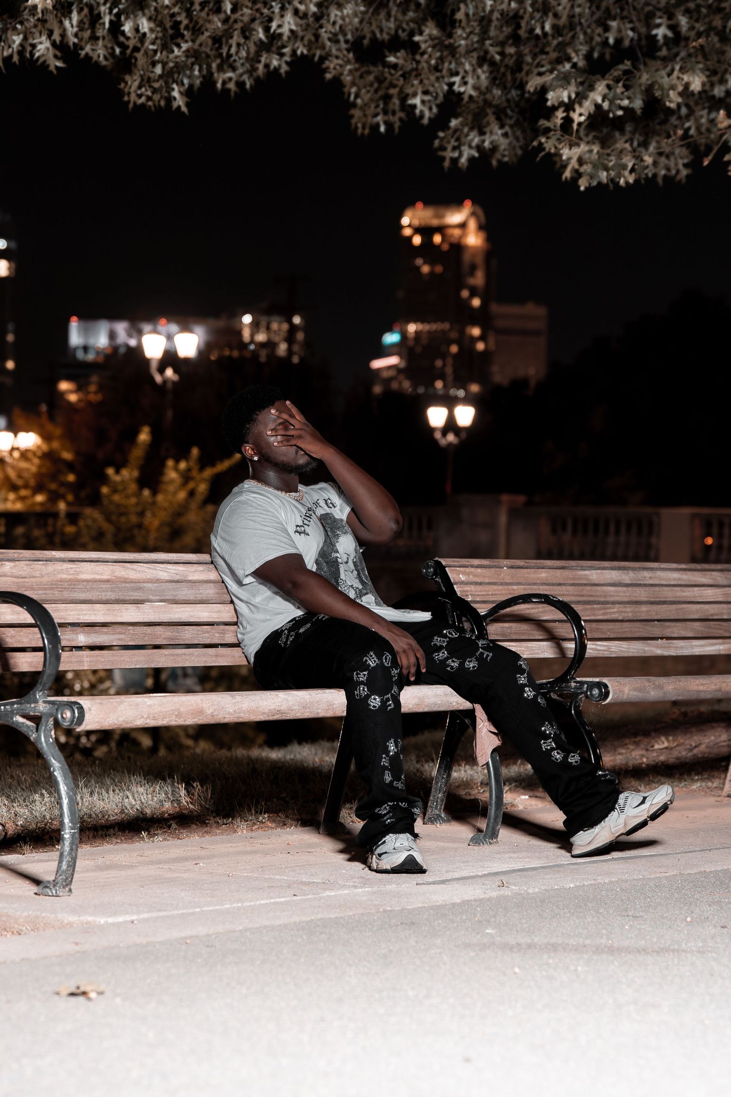 Young man relaxing on a bench at night, leaning back under trees with urban lights behind him.