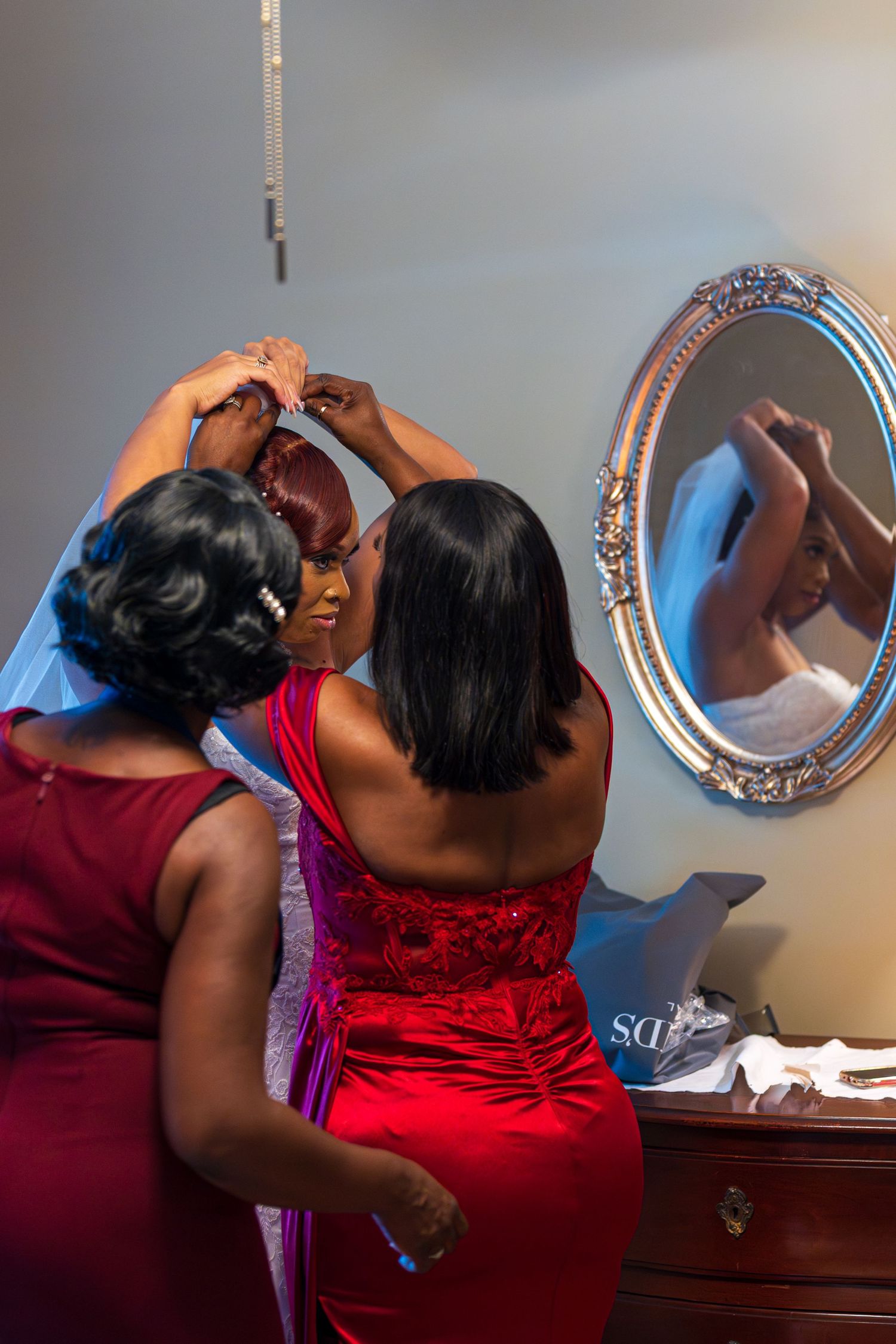 Bridesmaid in red dress helping bride with hair accessories reflected in an ornate silver mirror.