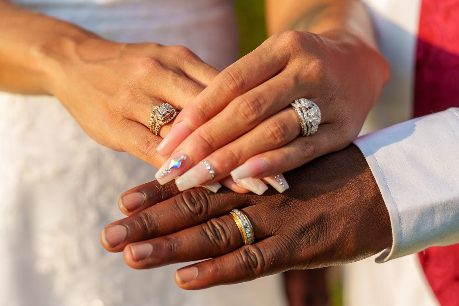 Interracial couple's hands stacked together displaying wedding rings at outdoor ceremony.