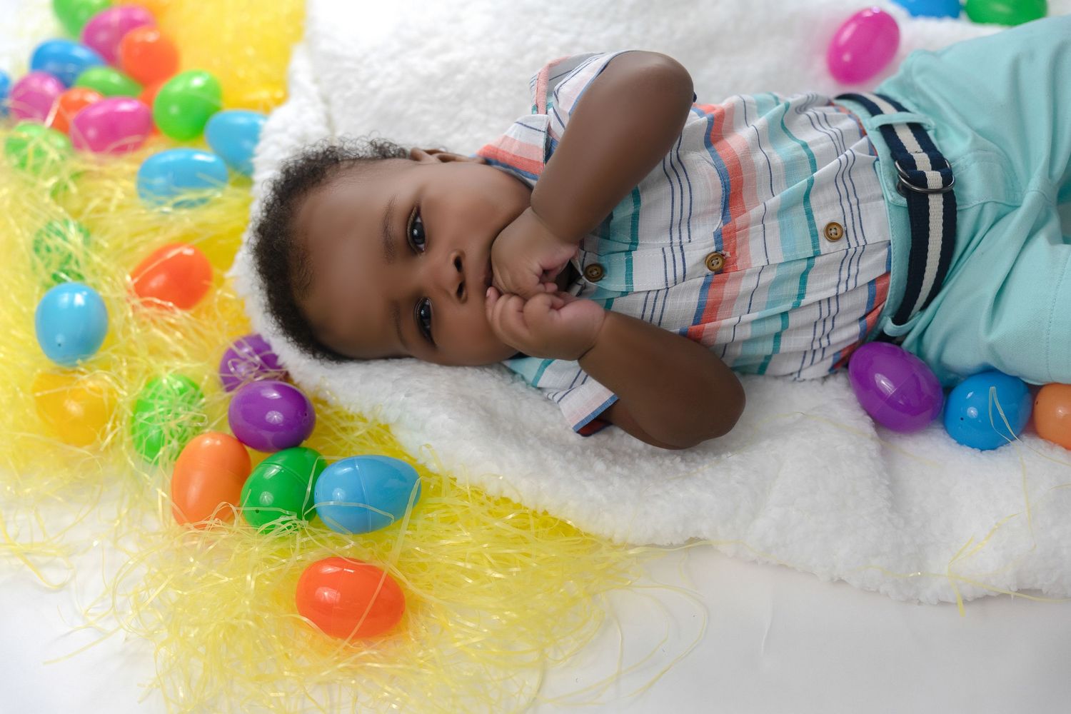Cute baby lying among colorful Easter eggs on a white fluffy blanket in a studio.