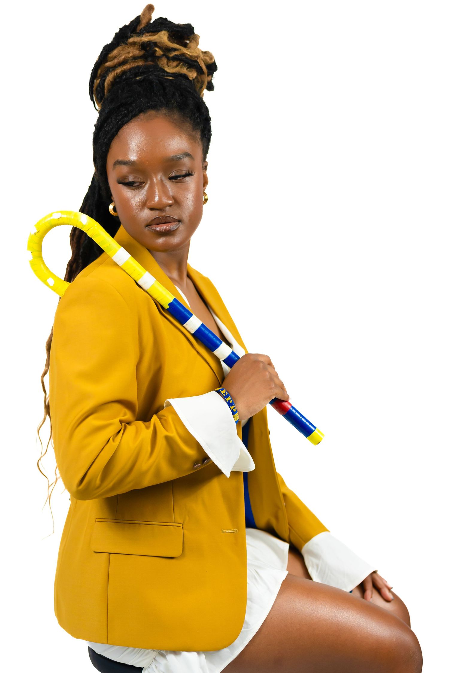Young Black woman in yellow blazer holding colorful cane prop in white studio setting.
