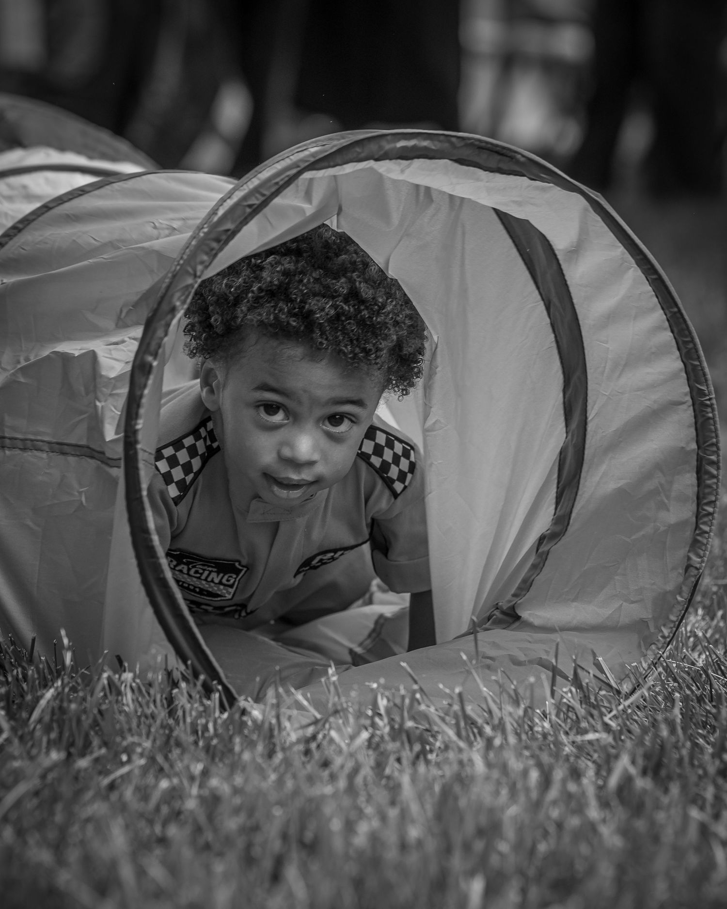 Black and white photo of a young boy crawling through a colorful play tunnel on grass.