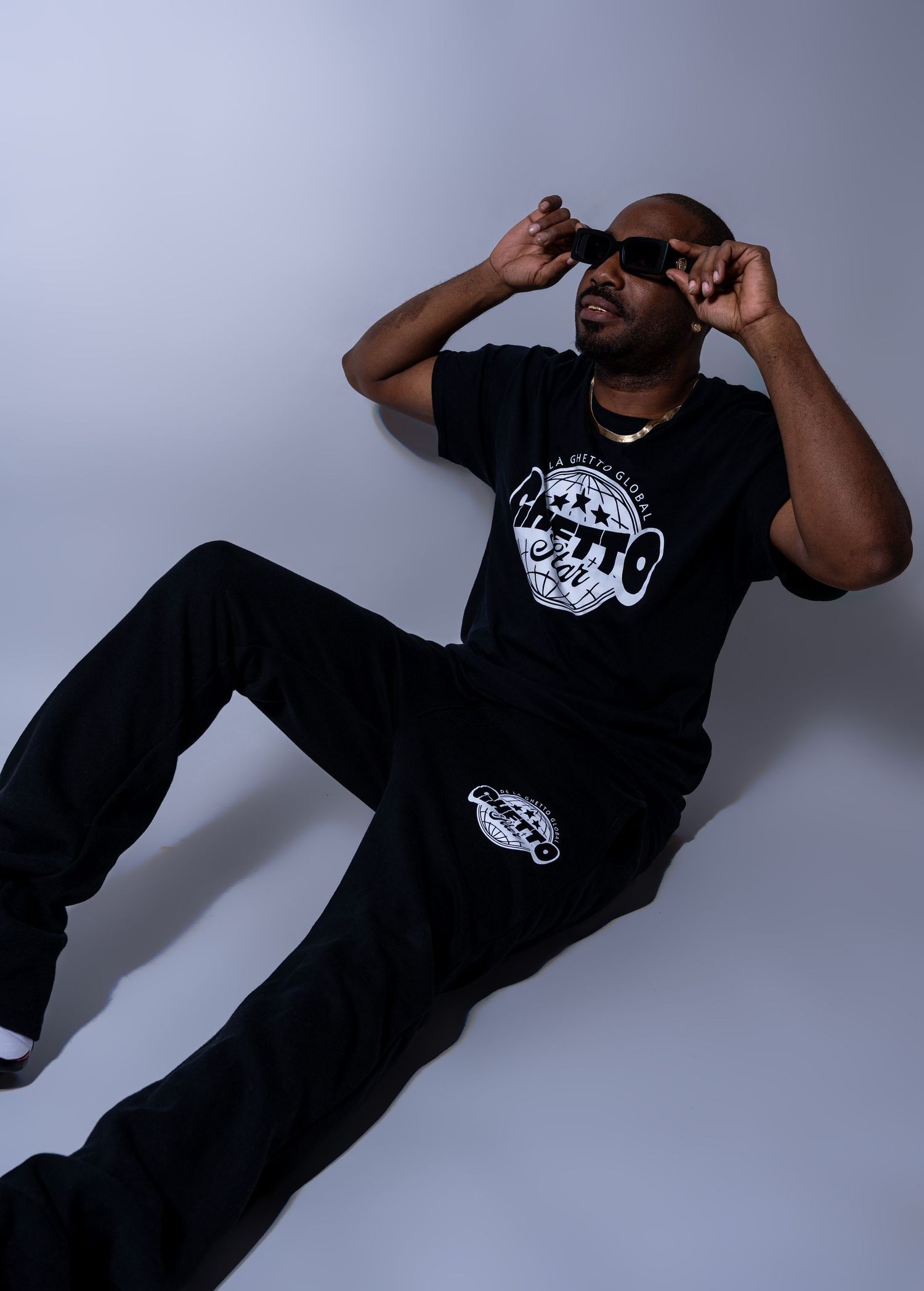 Man in black streetwear outfit lying on floor adjusting sunglasses in minimalist studio.
