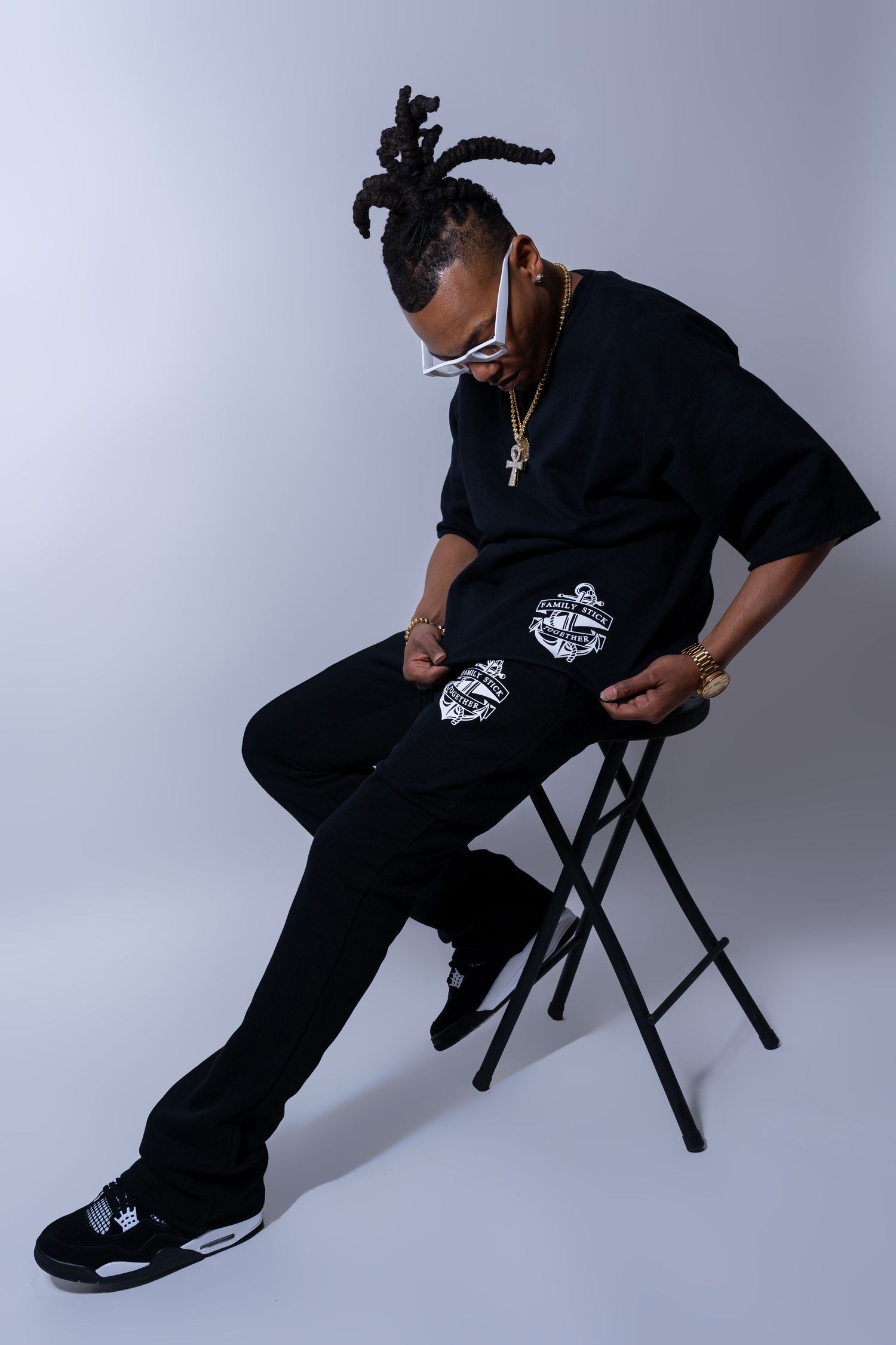 Man in black oversized tee and pants with white graphic patches, seated on stool.
