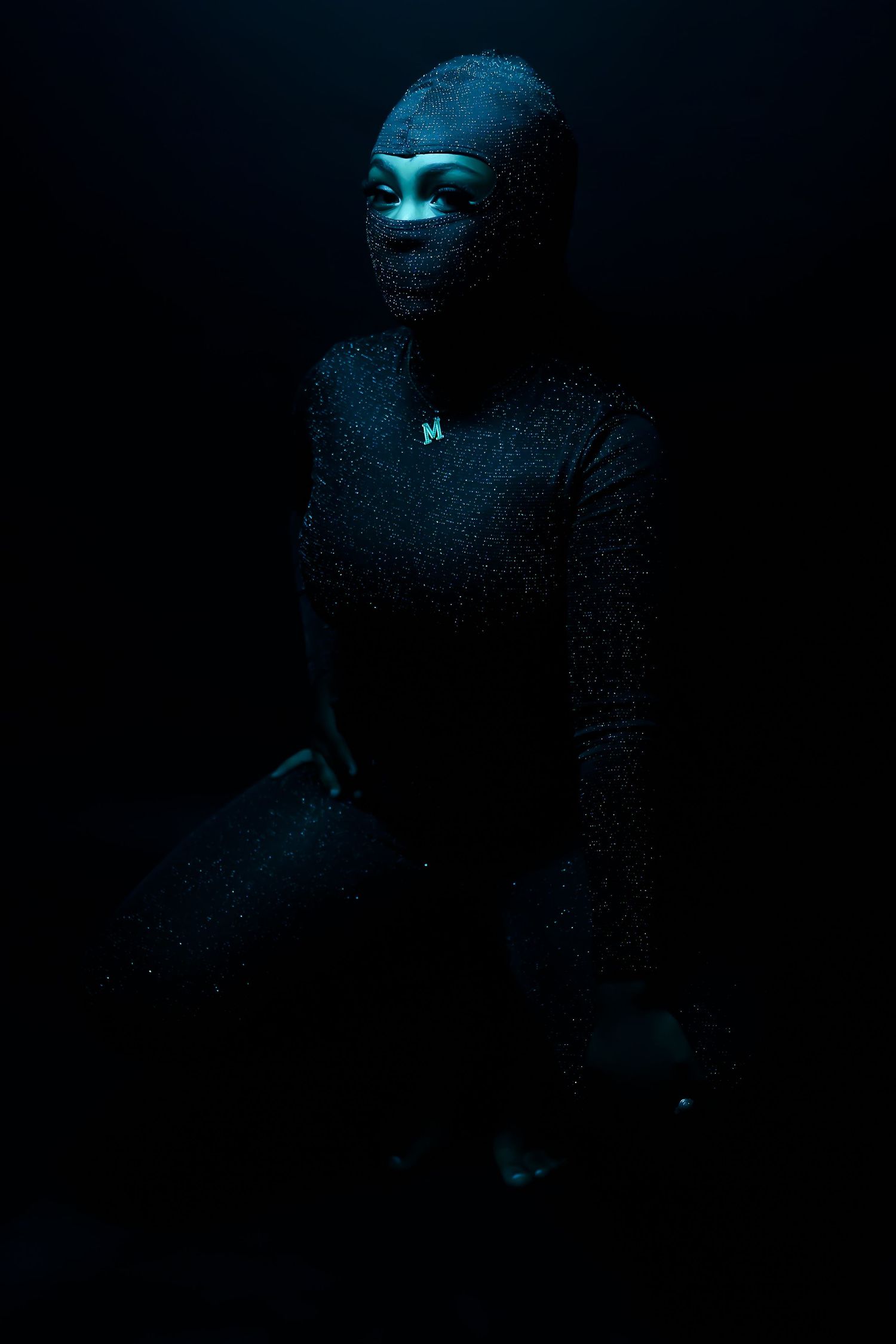 Person covered in dark glitter bodysuit kneeling in dramatic blue-tinted studio lighting against black background.