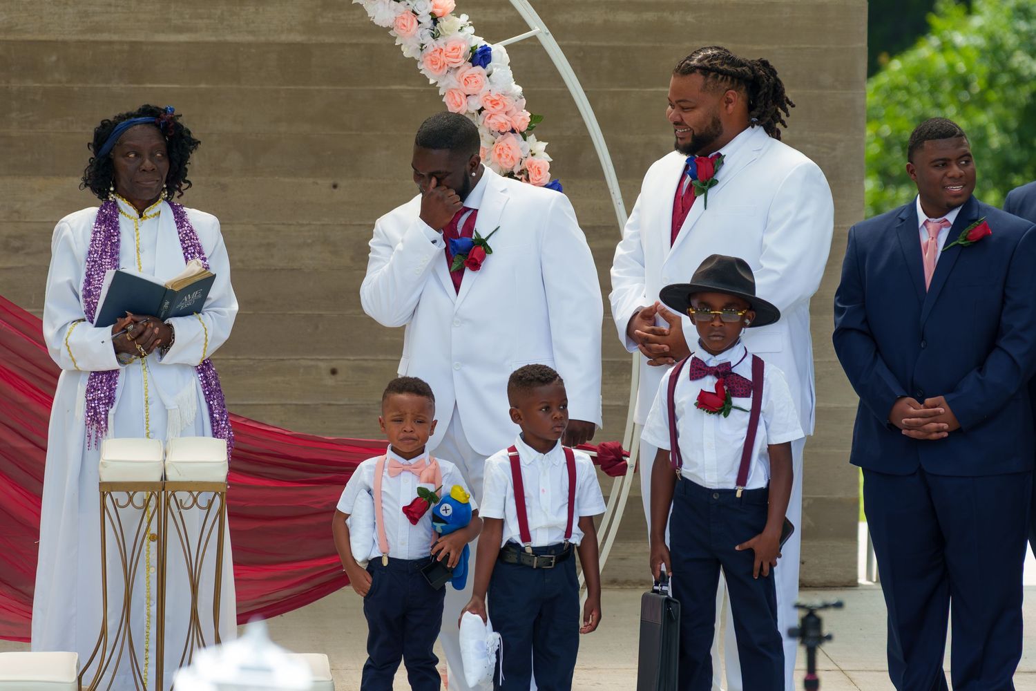Groom in white suit with groomsmen and children in suspenders at outdoor wedding ceremony.