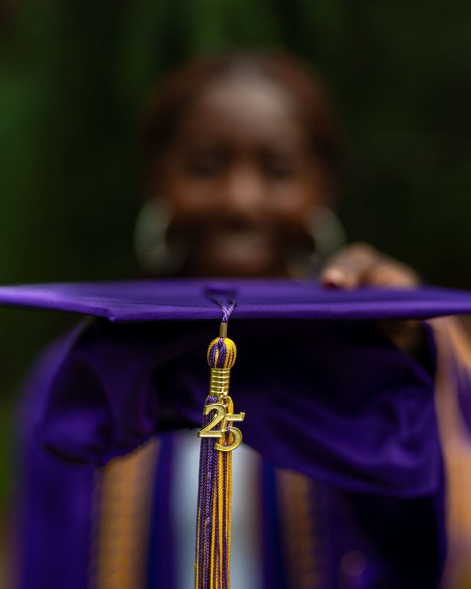 Close-up of purple graduation cap with gold 2025 tassel held up by blurred smiling graduate in background.