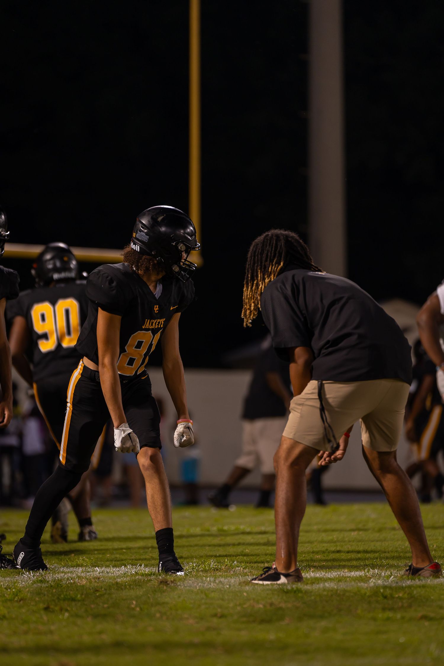High school football player number 81 in black jersey receives coaching instructions during night game.