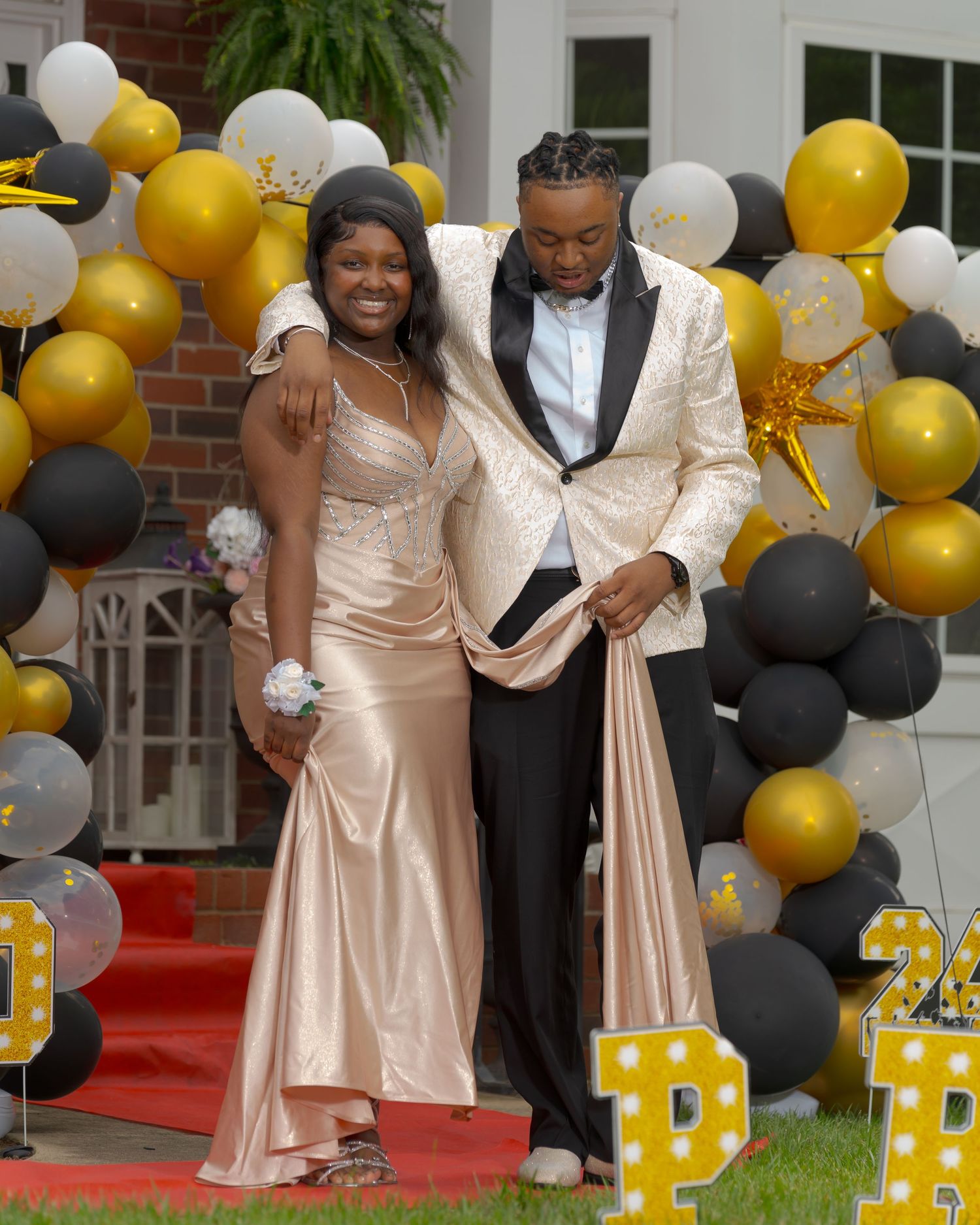 Prom couple posing before black gold and white balloon arch with PROM letter decorations on red carpet.
