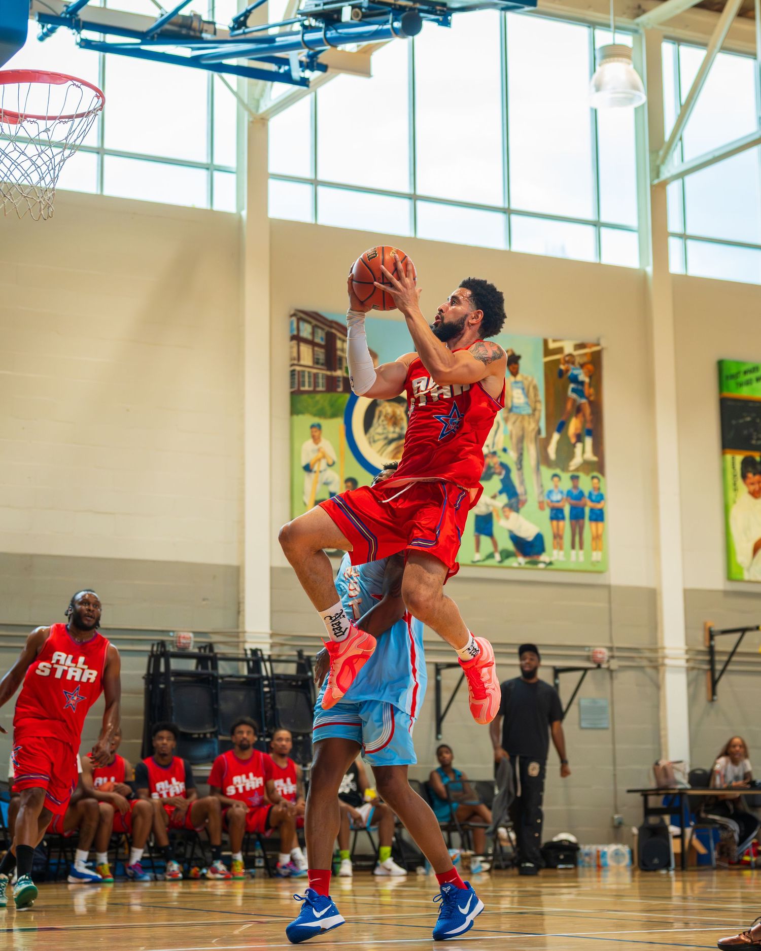 Basketball player in red All-Star jersey drives to basket for layup during indoor game.