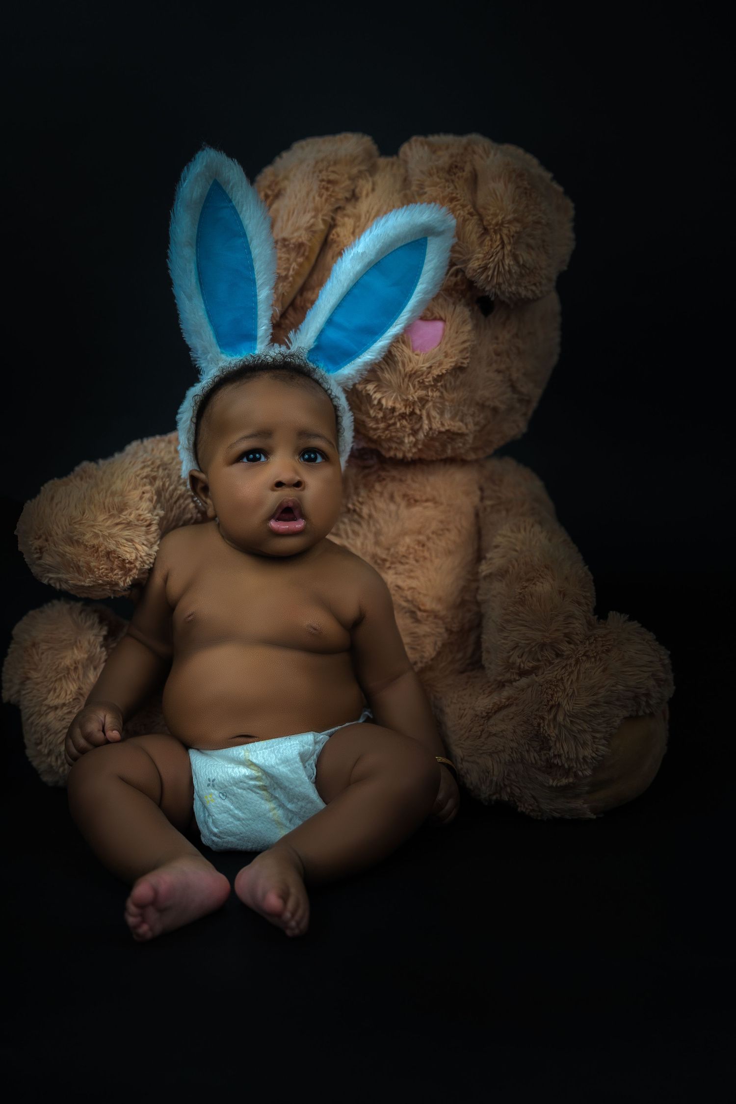 Baby in diaper with blue bunny ears sits against large teddy bear on black background.