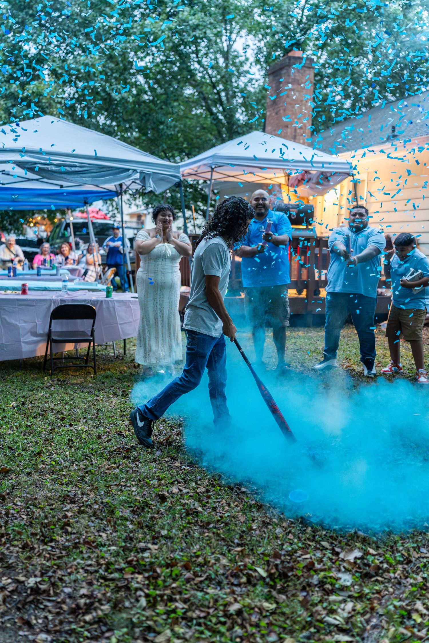 Person releasing blue smoke cannon at outdoor gender reveal party with excited guests and confetti.