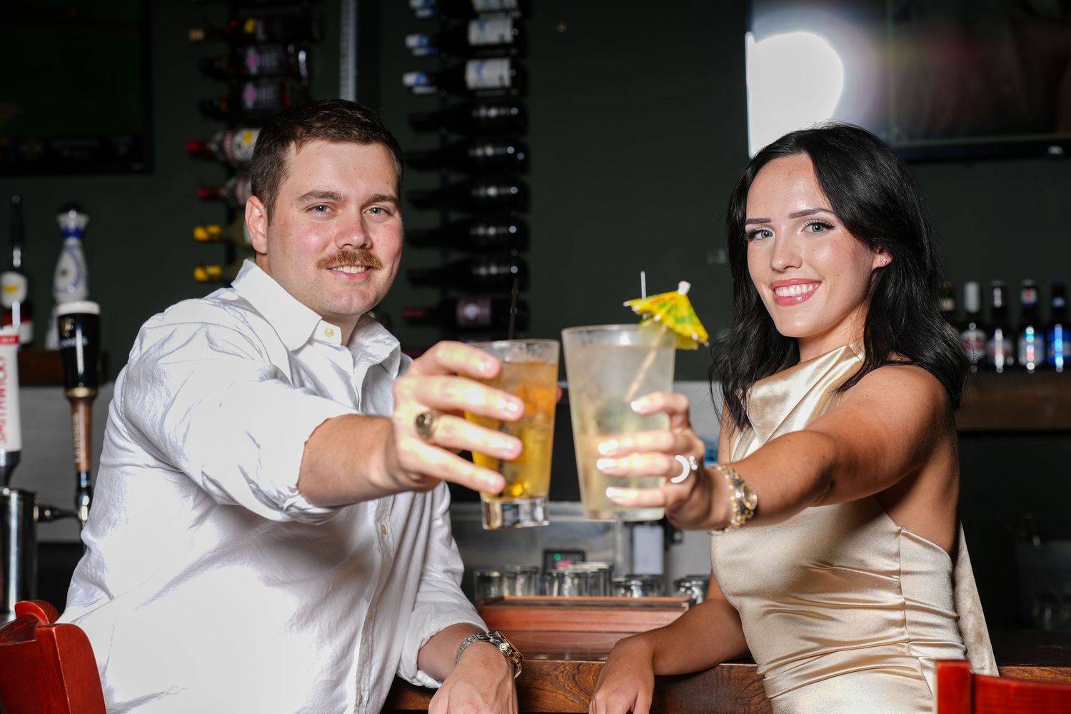 Couple toasting cocktails at a bar, woman in gold dress and man in white shirt smiling at camera.