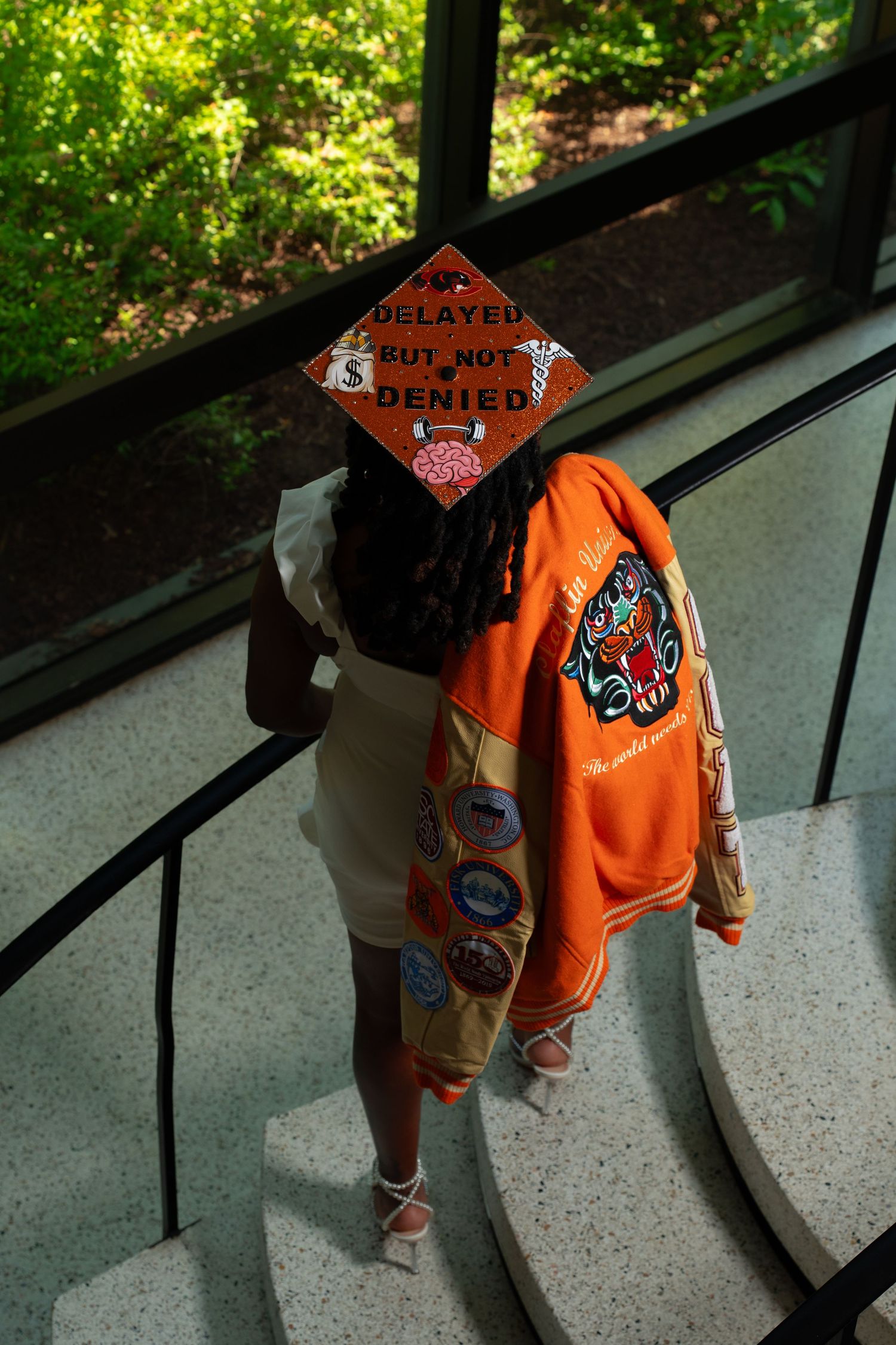 Graduate wearing decorated mortarboard reading Delayed But Not Denied with orange jacket on spiral staircase.