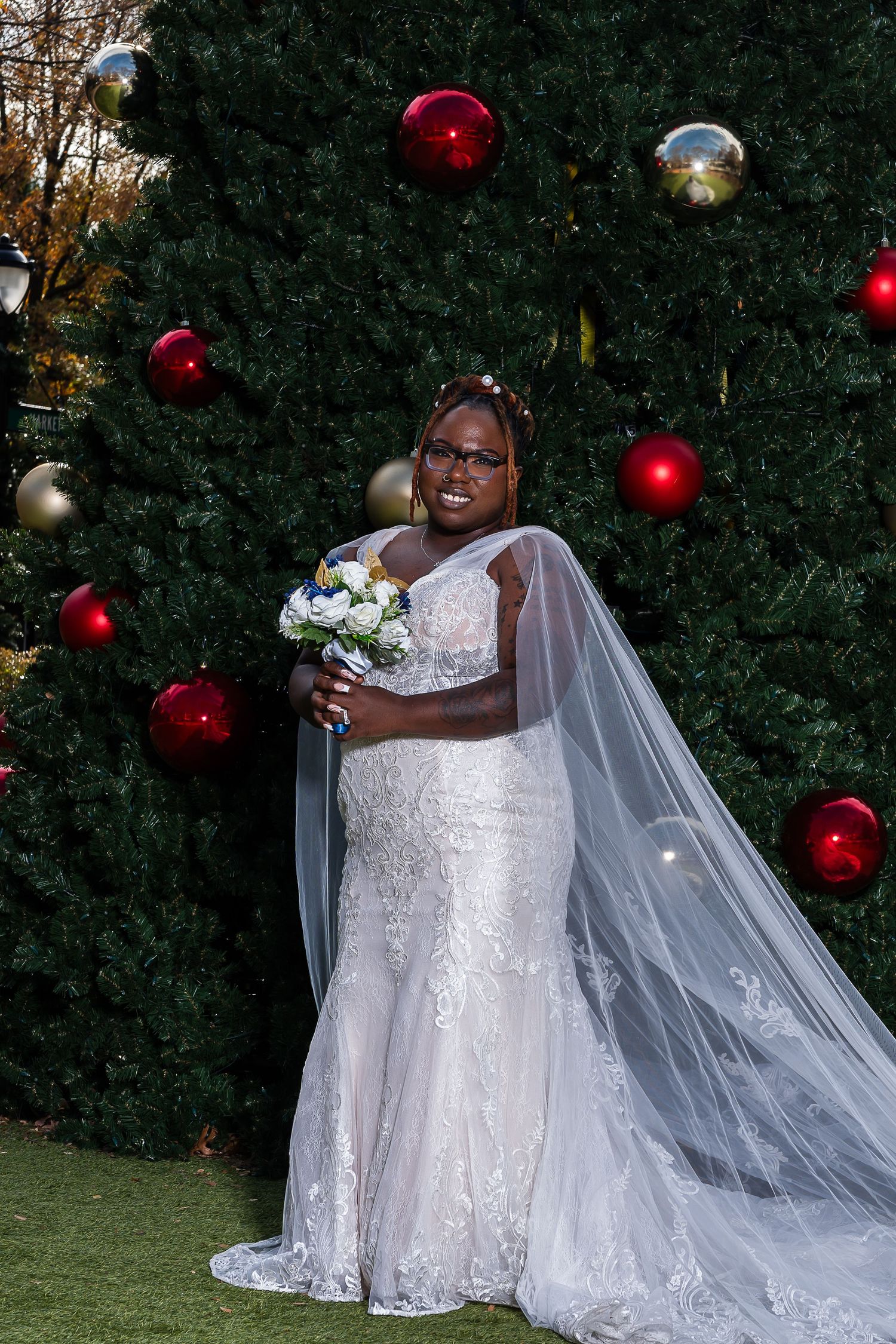 Bride in white lace gown with cape veil holds bouquet before decorated Christmas tree with red ornaments.