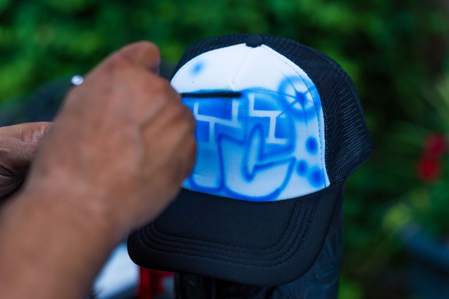 Close-up of a black and white trucker hat with blue airbrush graffiti lettering being held outdoors.