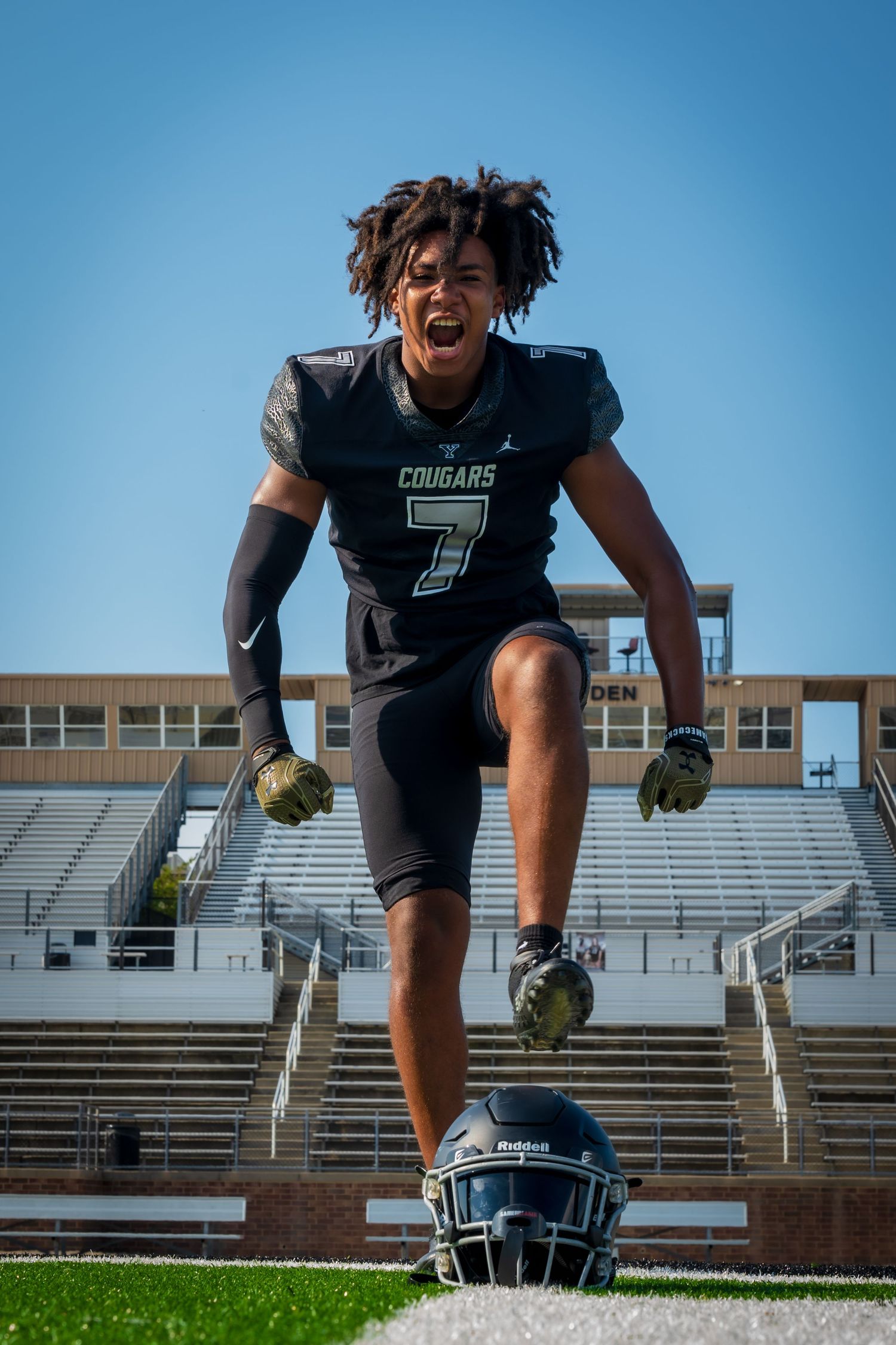 Football player #7 in black Cougars jersey poses aggressively on field with helmet at feet.
