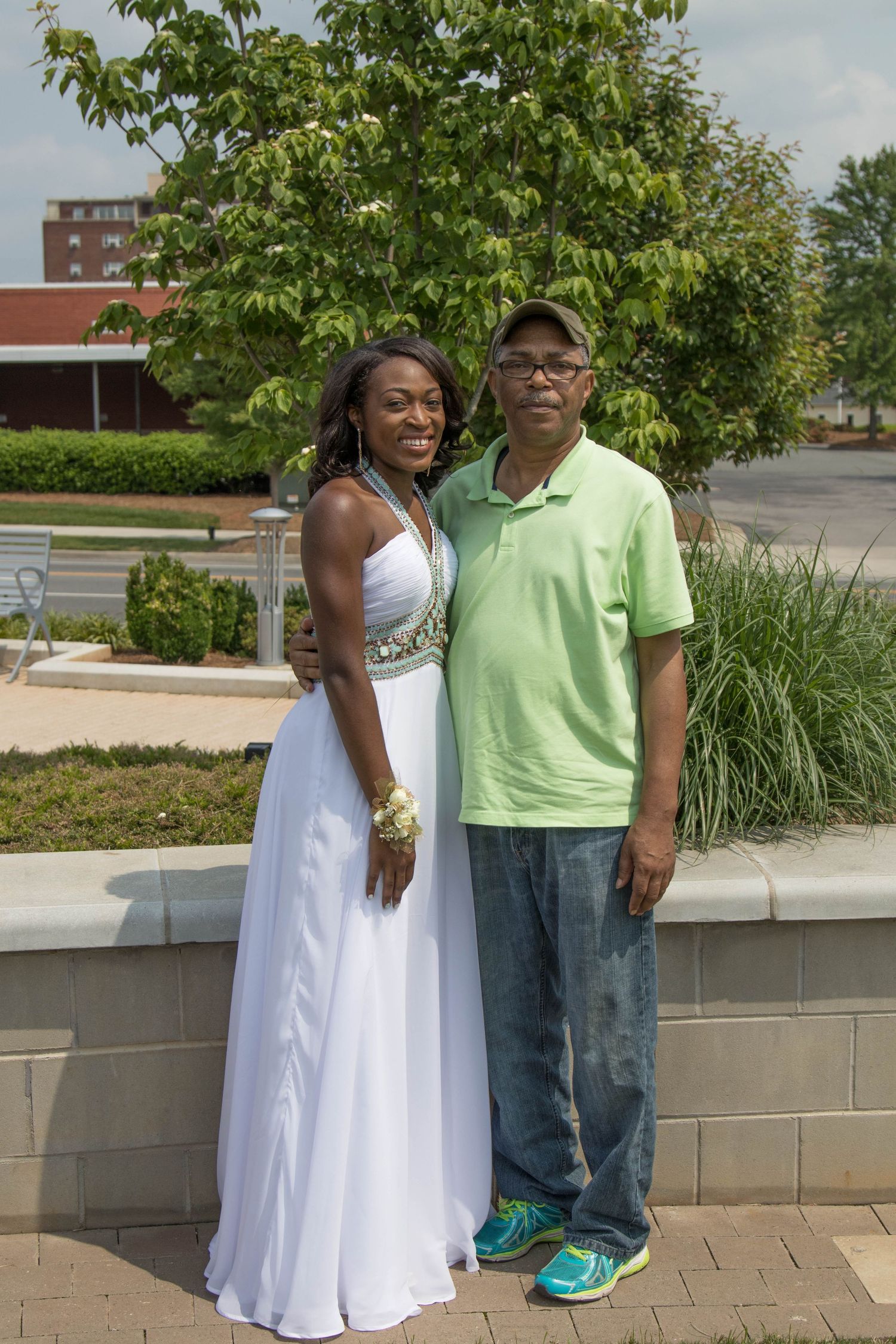 A young woman in a white prom gown and a man in a green polo shirt pose together outdoors near trees.