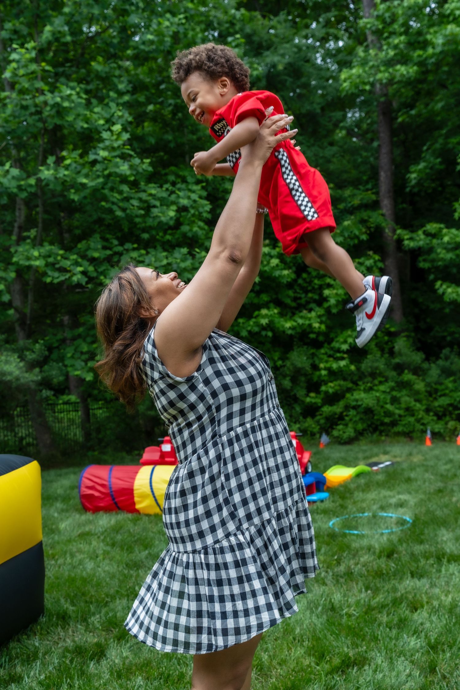 Woman in checkered dress lifts smiling toddler in red racing outfit high in the air outdoors.