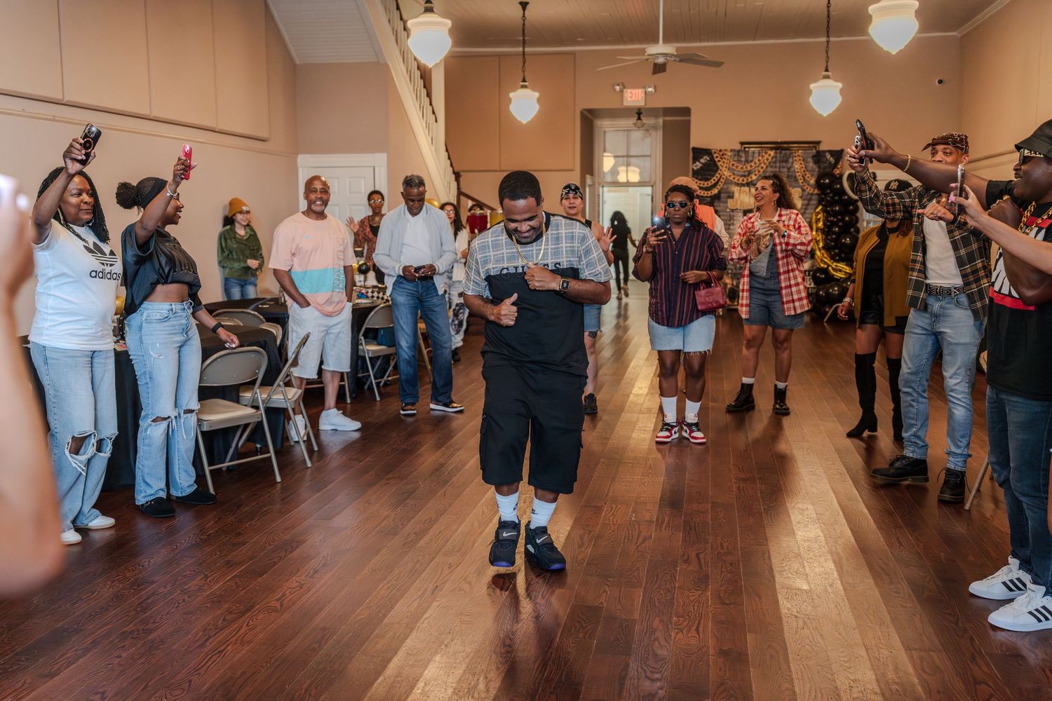 Young man dancing in center of cheering crowd on wooden dance floor at an indoor celebration.