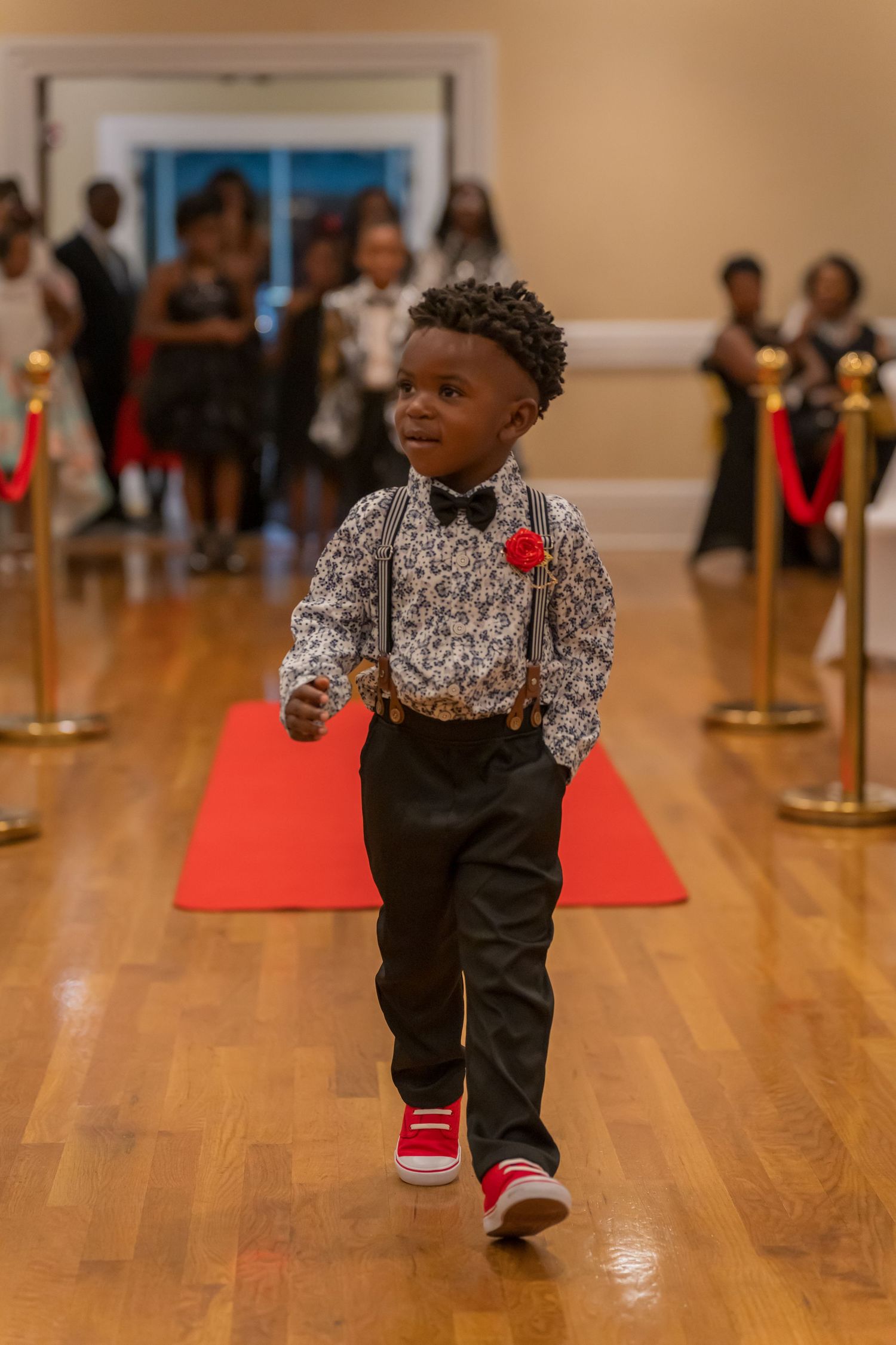 A young boy in suspenders and a bow tie walks a red carpet at a formal gala event.