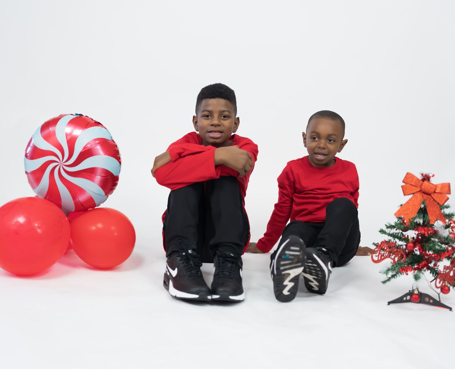 Two boys in red shirts sit together beside red balloons and a small decorated Christmas tree.