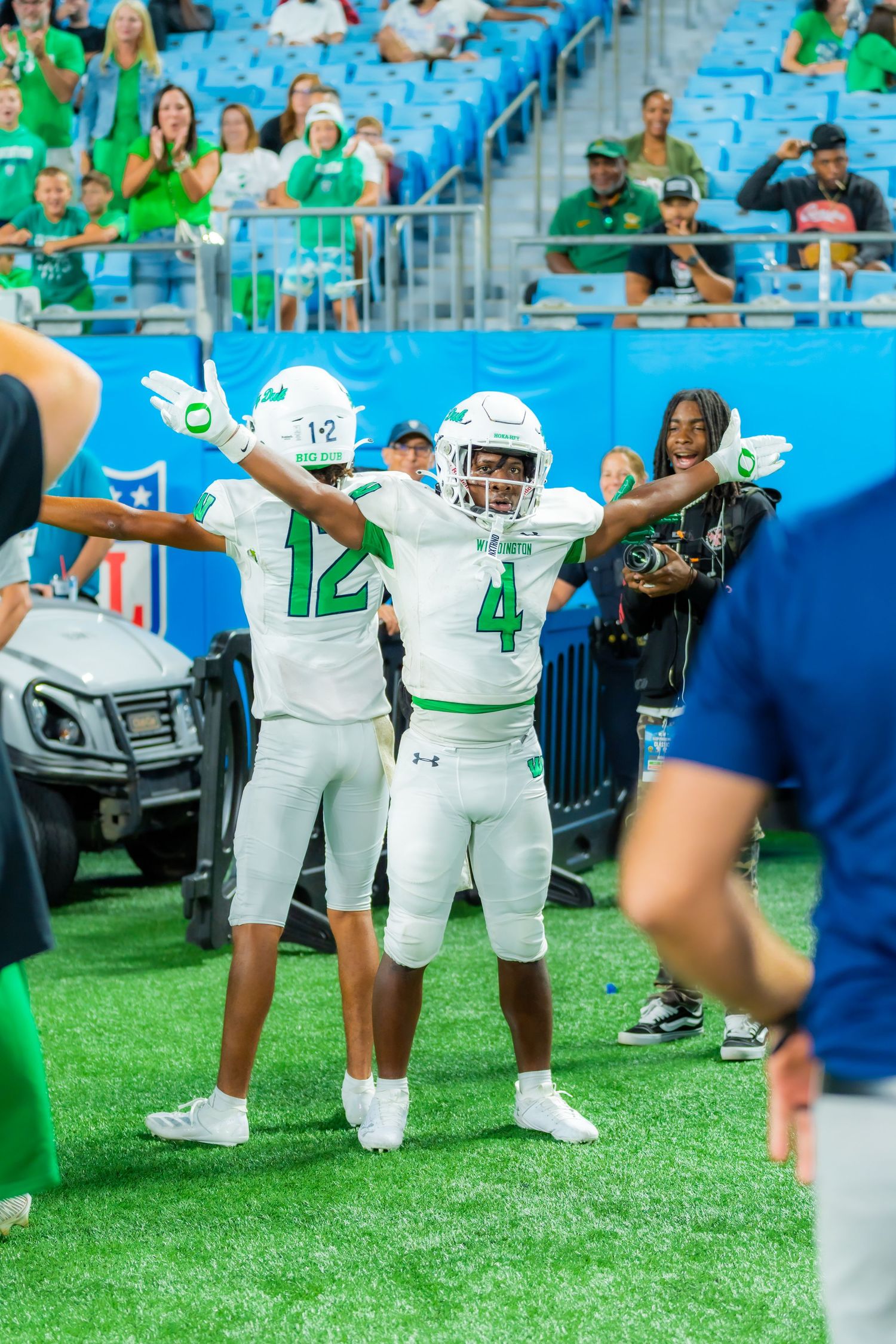 Two football players in green uniforms numbers 12 and 4 celebrate on stadium field.