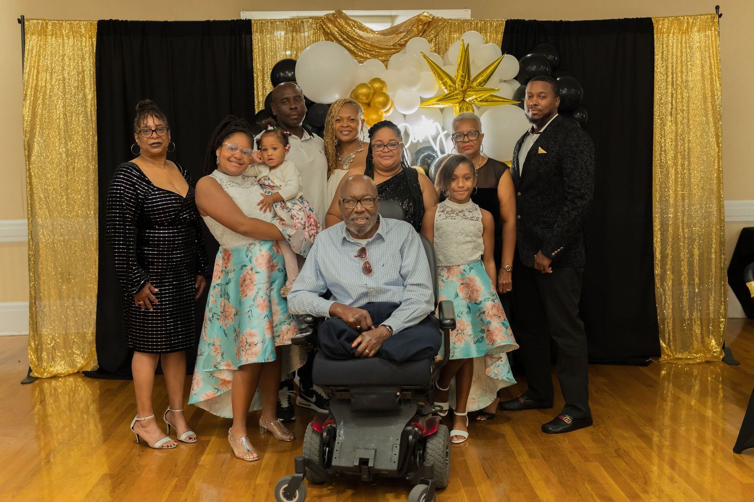 Family group photo at a celebration with gold balloons, including a man in a motorized wheelchair.