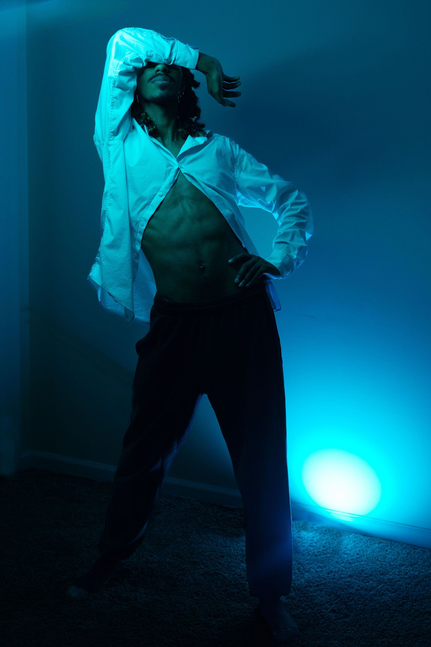 Man posing in open white shirt under blue light indoors.