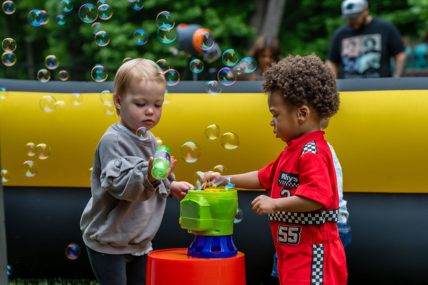 Two toddlers play with a bubble machine outdoors, surrounded by floating bubbles at a party.