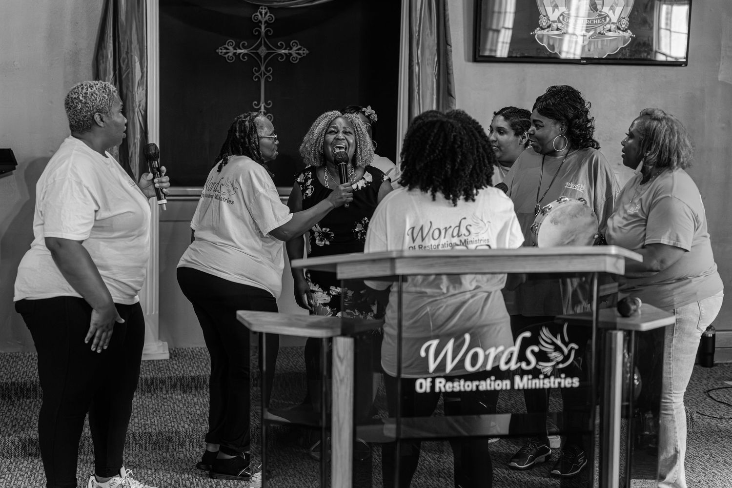 A black and white photo of women gathered around a Words of Restoration Ministries podium.