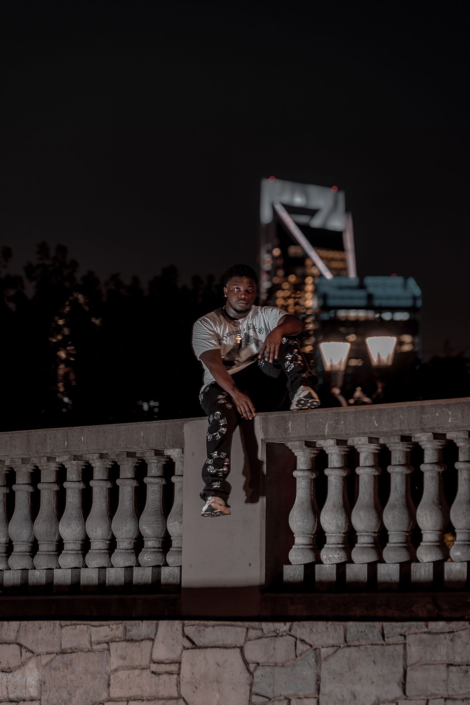 Man in white tee and camo pants sits on stone balustrade at night with city skyline behind him.