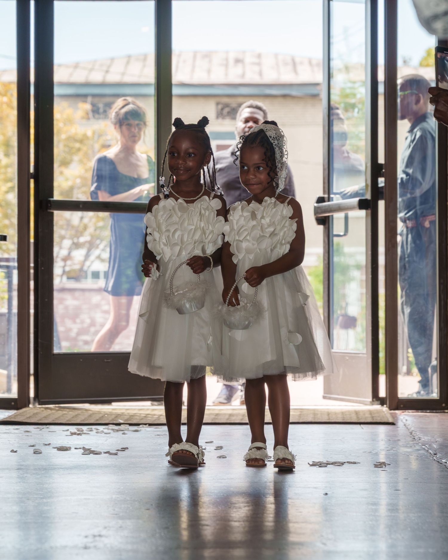 Two young flower girls in white dresses with floral details stand at ceremony entrance holding baskets.