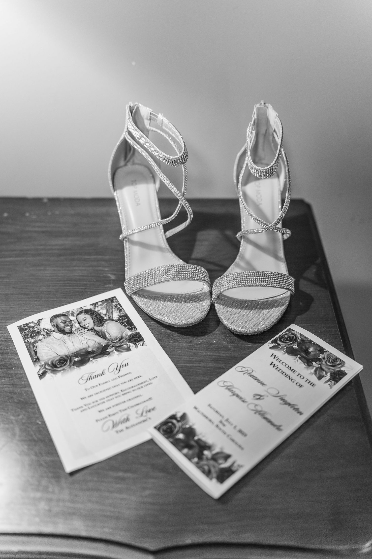 Black and white photo of glittery bridal heels beside wedding invitations on a wooden table.