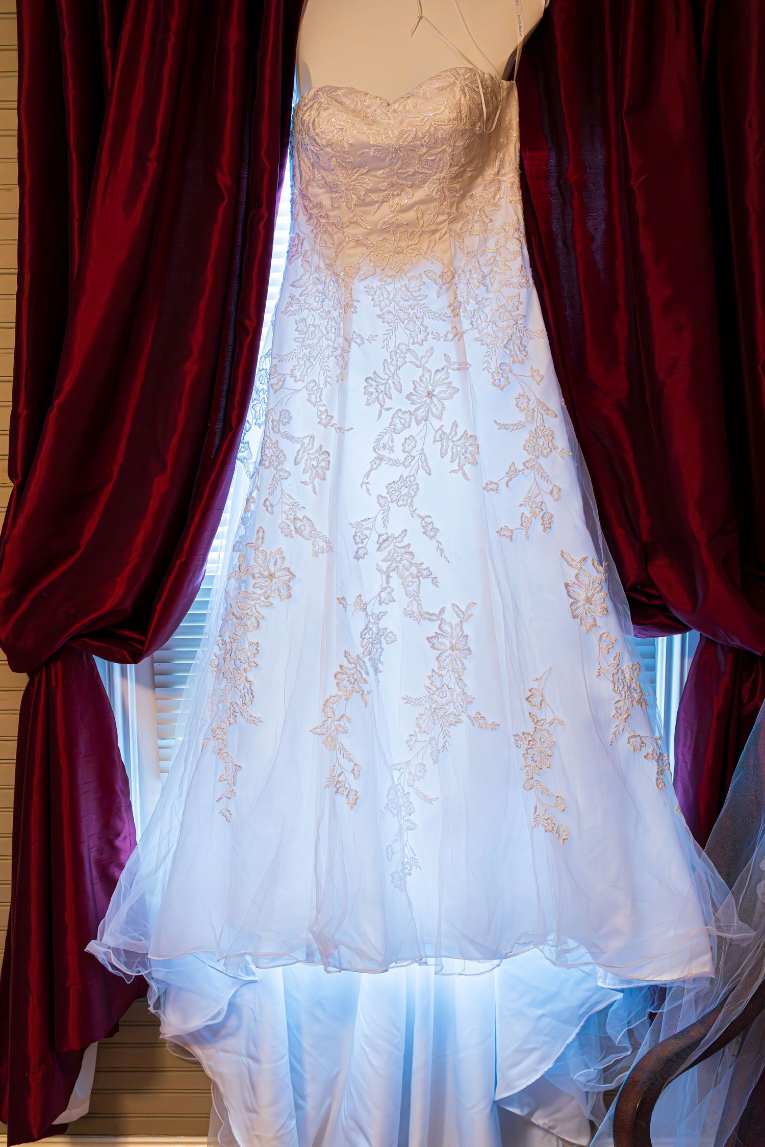 White lace wedding gown hanging between burgundy velvet curtains in window light.