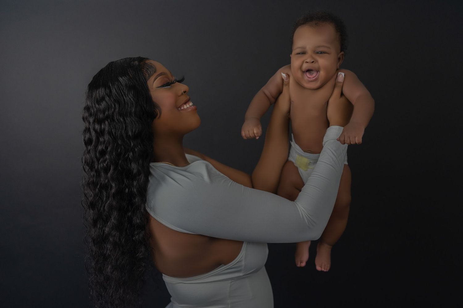 Laughing mother in white outfit holds giggling baby aloft against dark studio background.