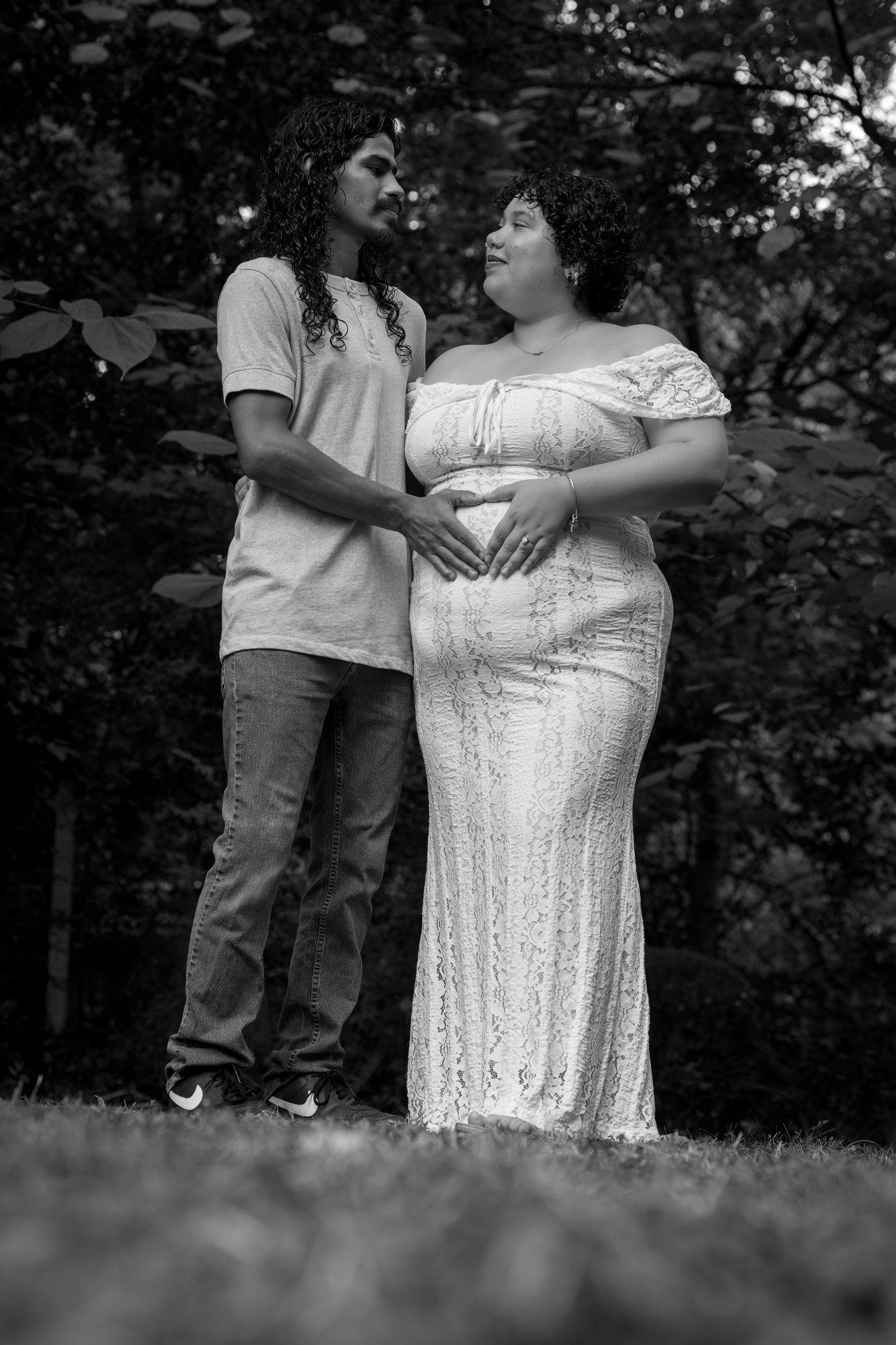 Black and white maternity photo of couple outdoors with woman in lace gown holding belly.
