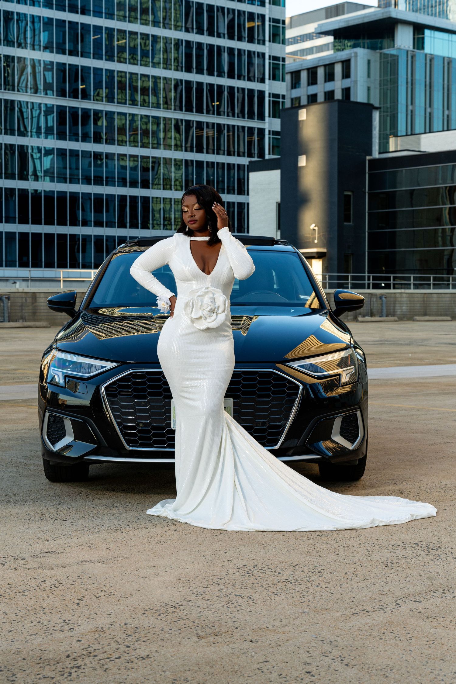 Young woman in white long-sleeve mermaid gown with train posing before black Audi in city.