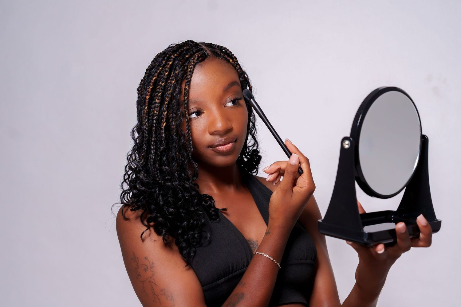 A young woman with braids applies makeup while holding a mirror against a white background.