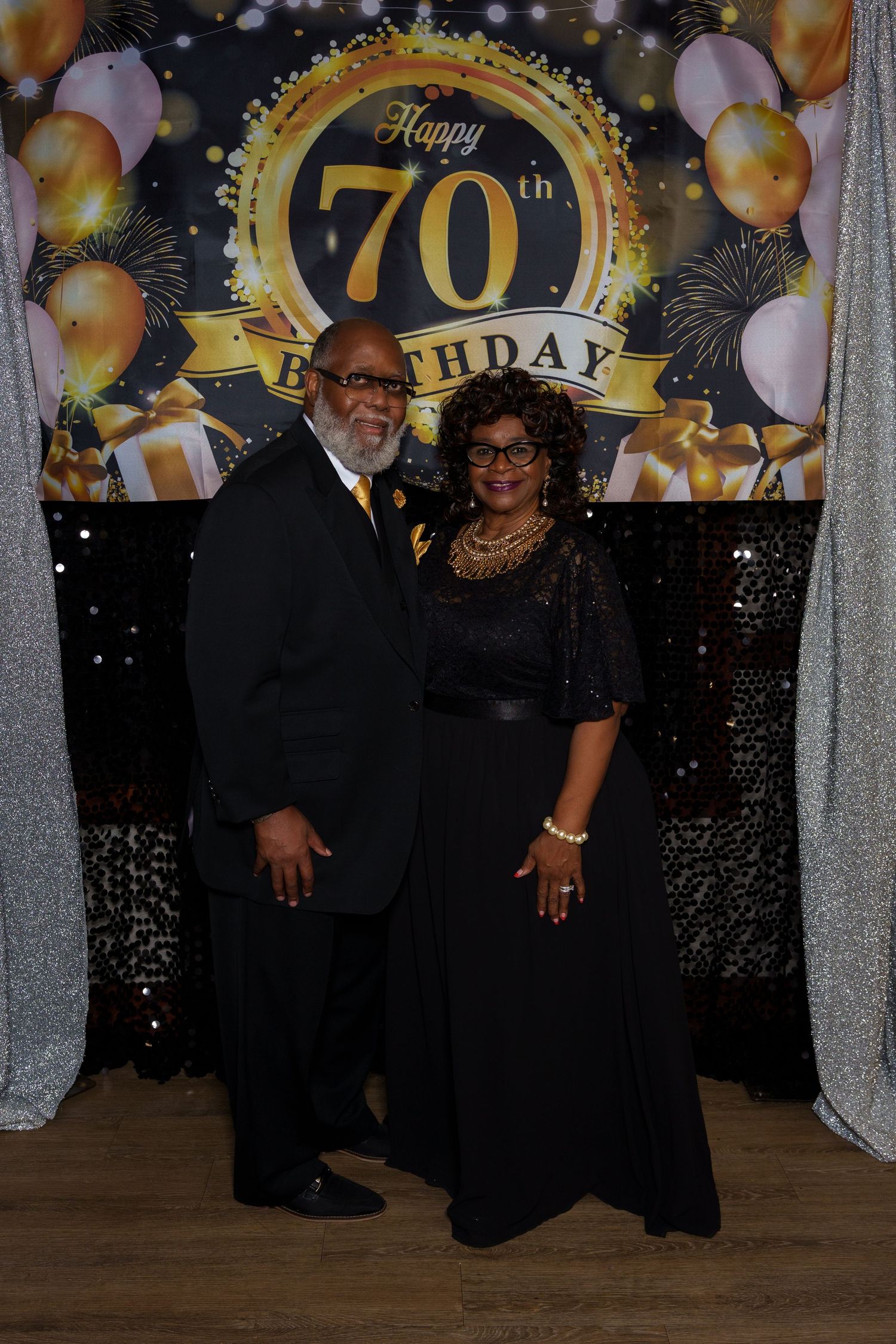 Elegant couple in black formal attire posing in front of a Happy 70th Birthday gold and black backdrop.