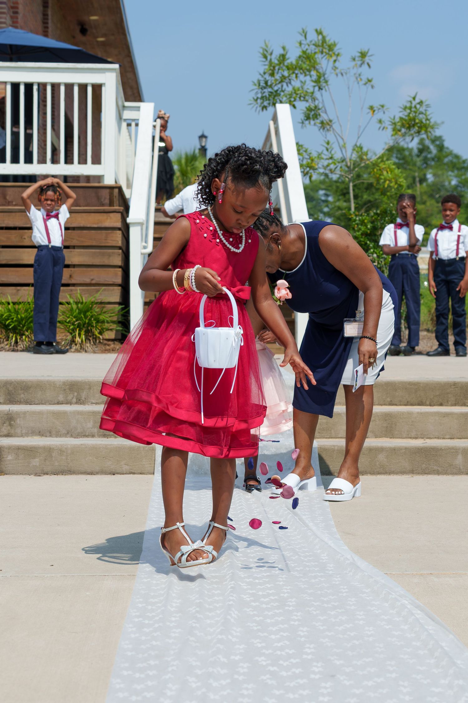 Flower girl in red dress scattering petals on white wedding aisle runner outdoors.