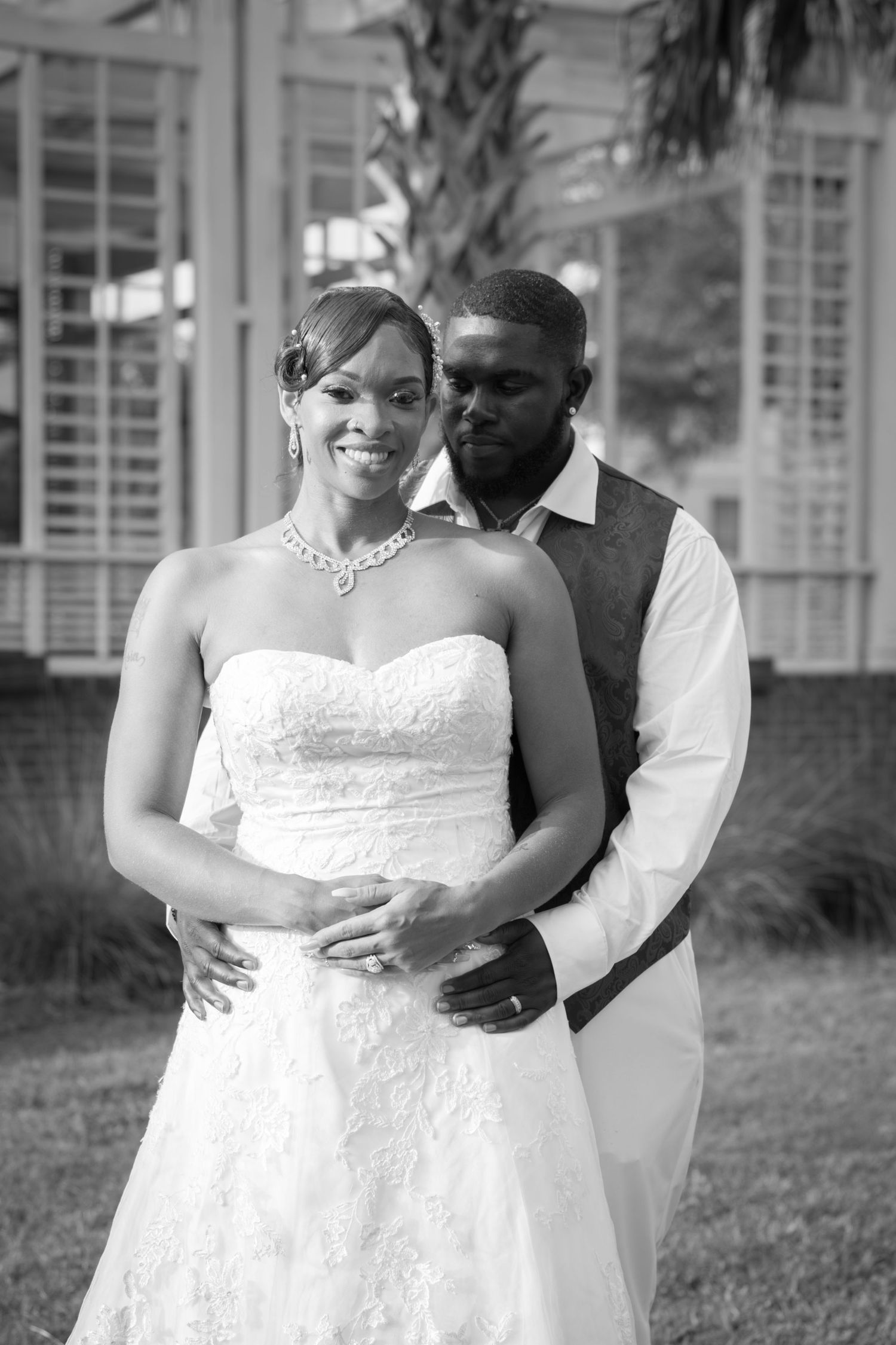 Black and white portrait of smiling bride in strapless gown embraced by groom outdoors.