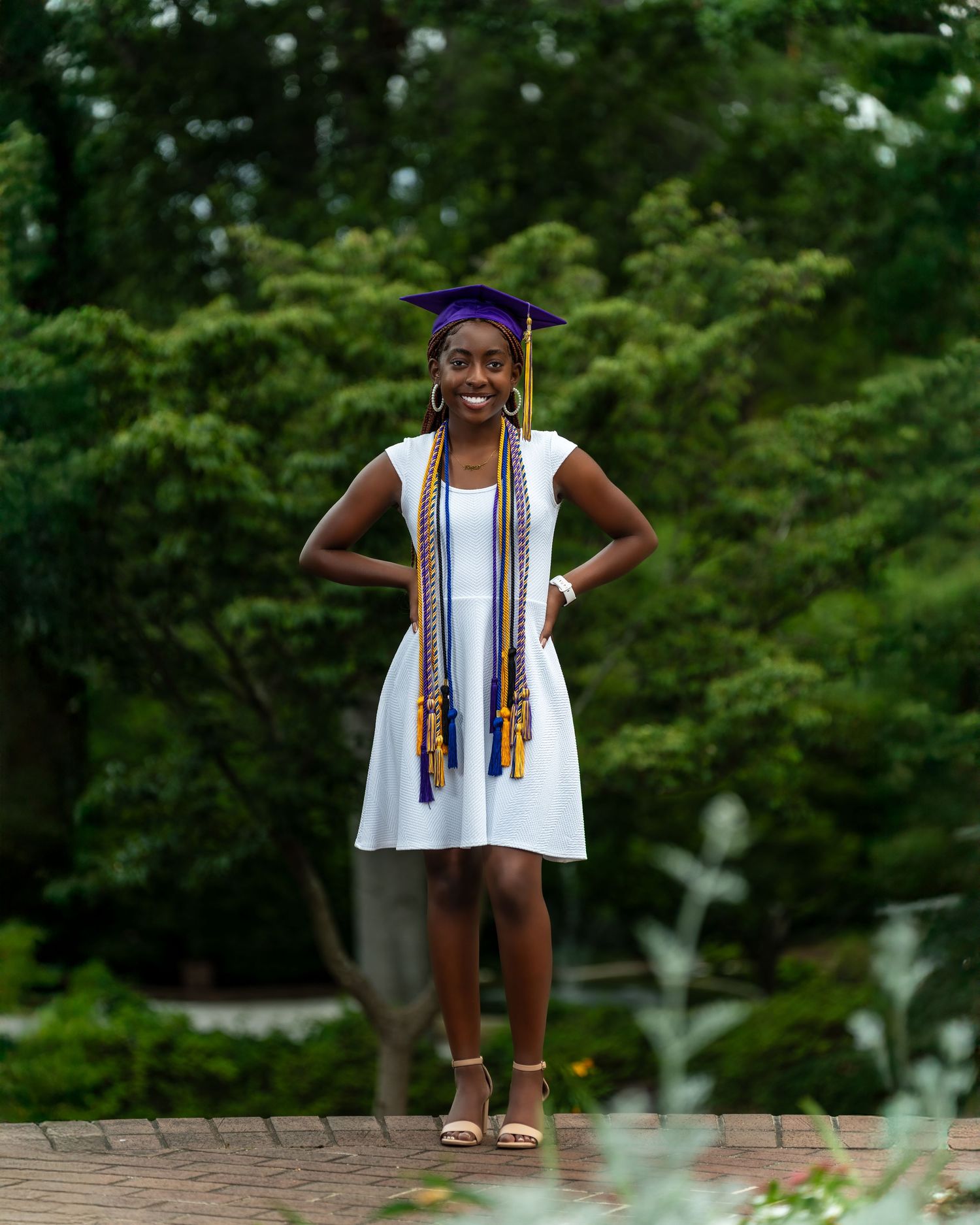 Smiling Black female graduate in white dress and purple cap with honor cords standing in lush green garden.