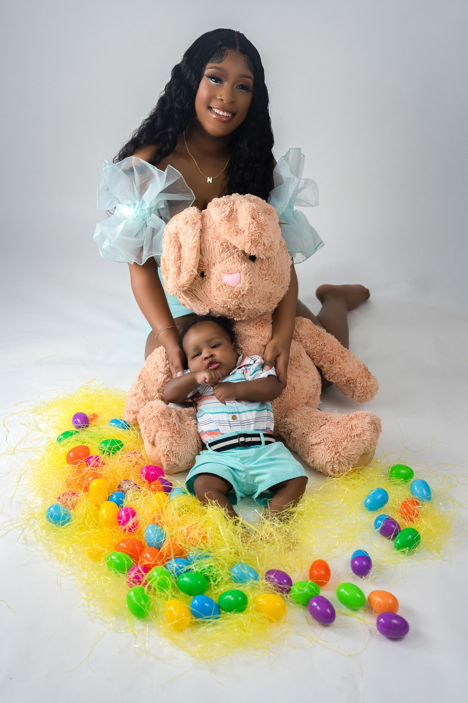 Woman and baby posed with a large Easter bunny stuffed animal surrounded by colorful Easter eggs.