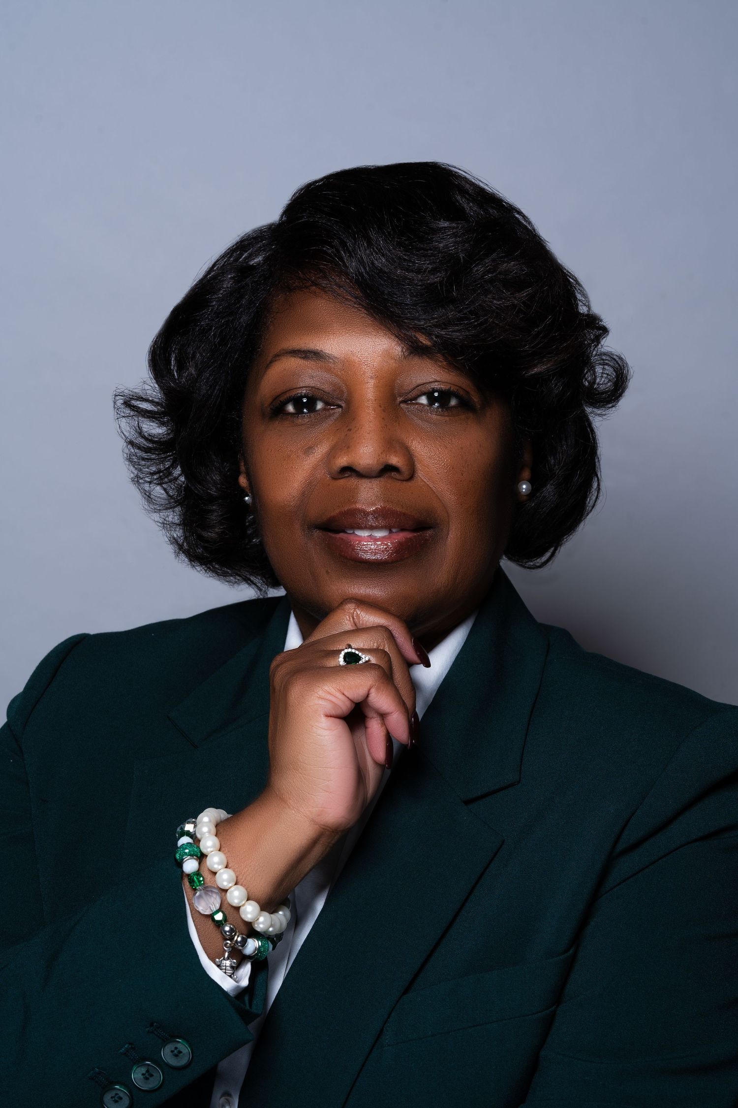 Professional Black woman in green blazer posing with hand on chin wearing pearl bracelet.