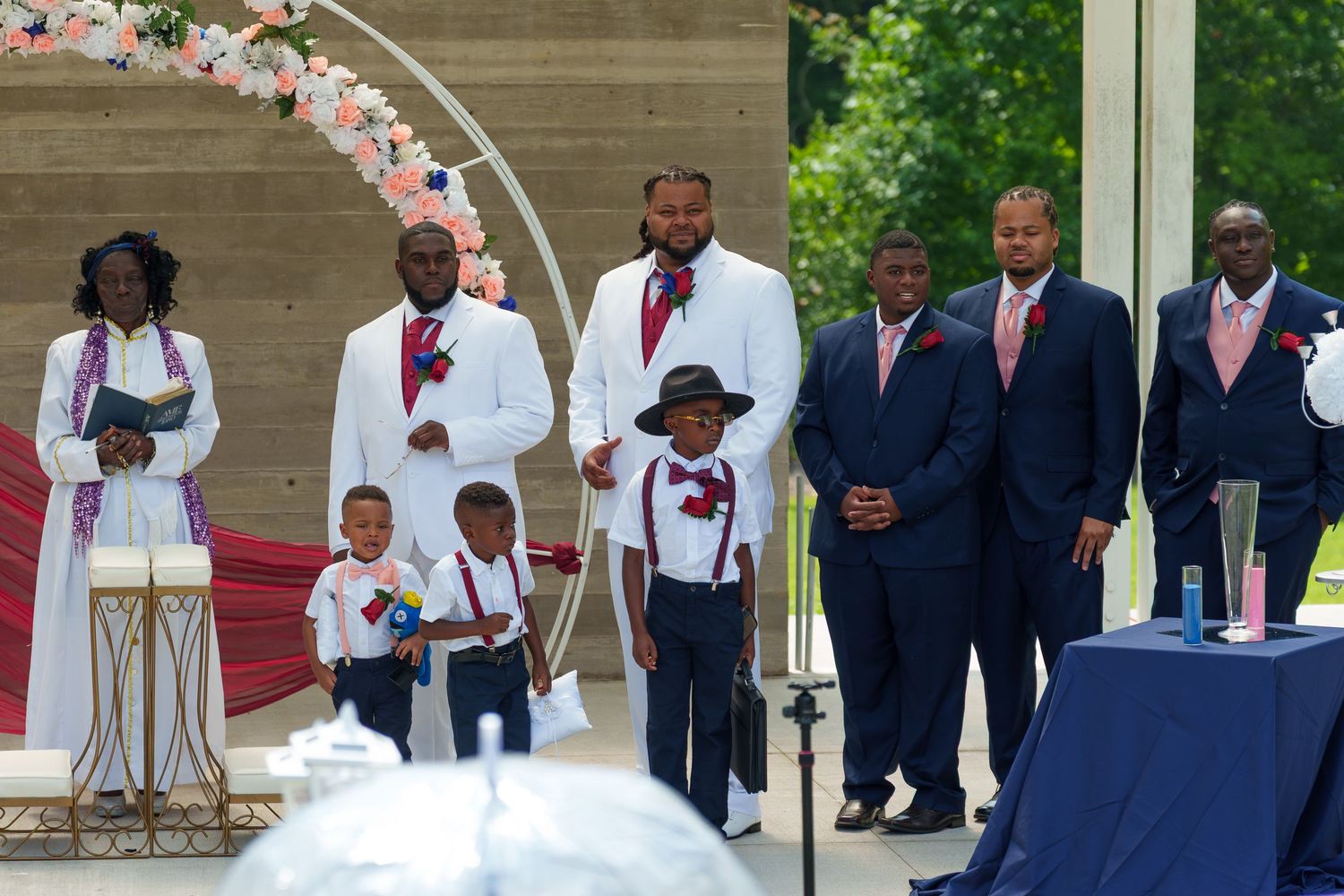 Wedding party in white and navy suits with children at outdoor ceremony under floral arch.