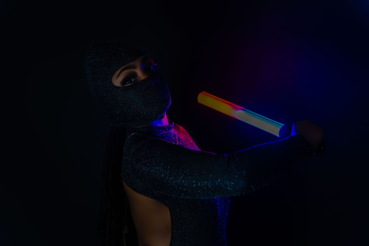 Model in glittery bodysuit holding colorful neon light tube in dark studio with purple and pink lighting.