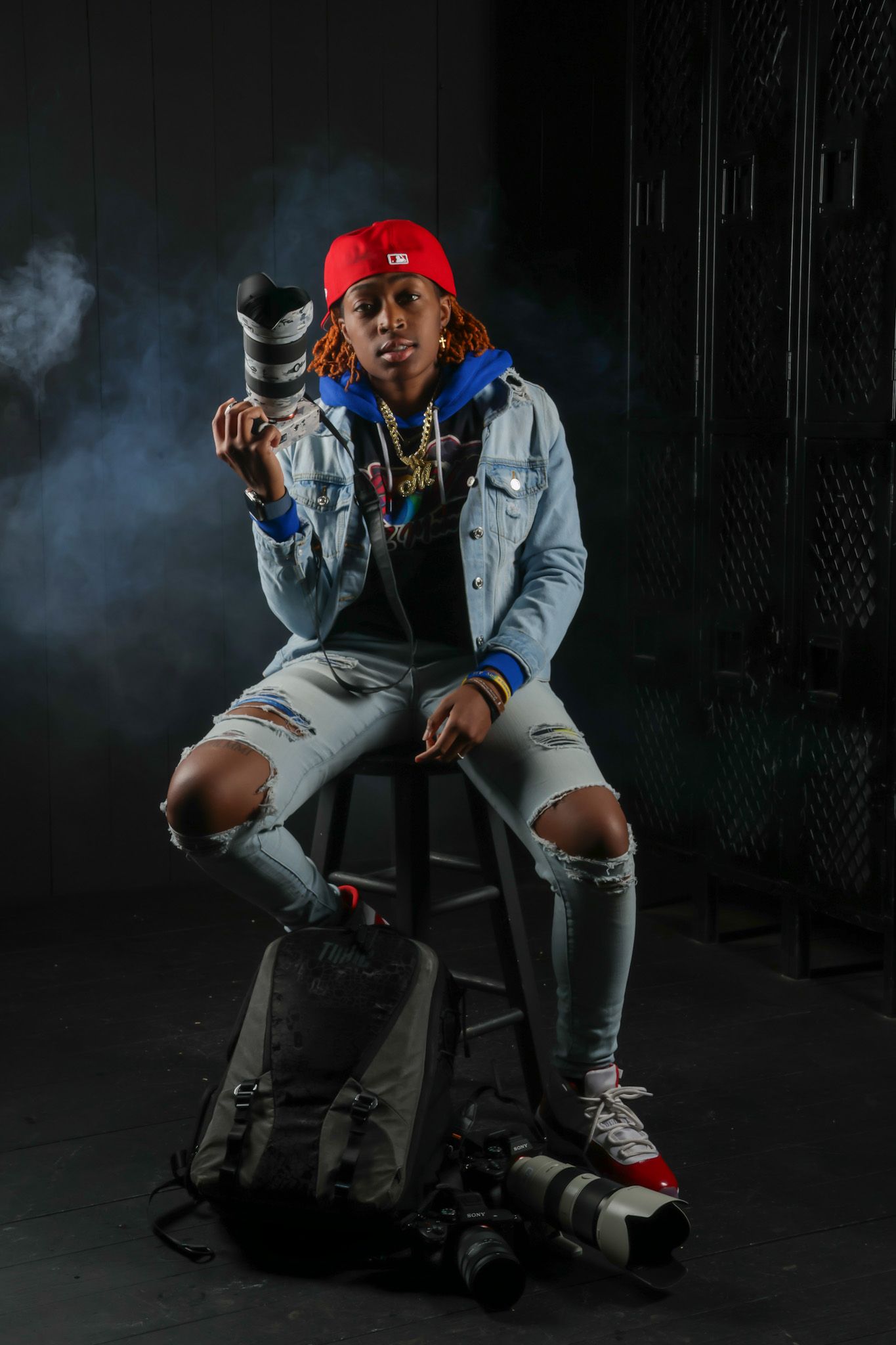 Young photographer sitting on stool holding camera lens wearing red cap and ripped jeans in dark studio.