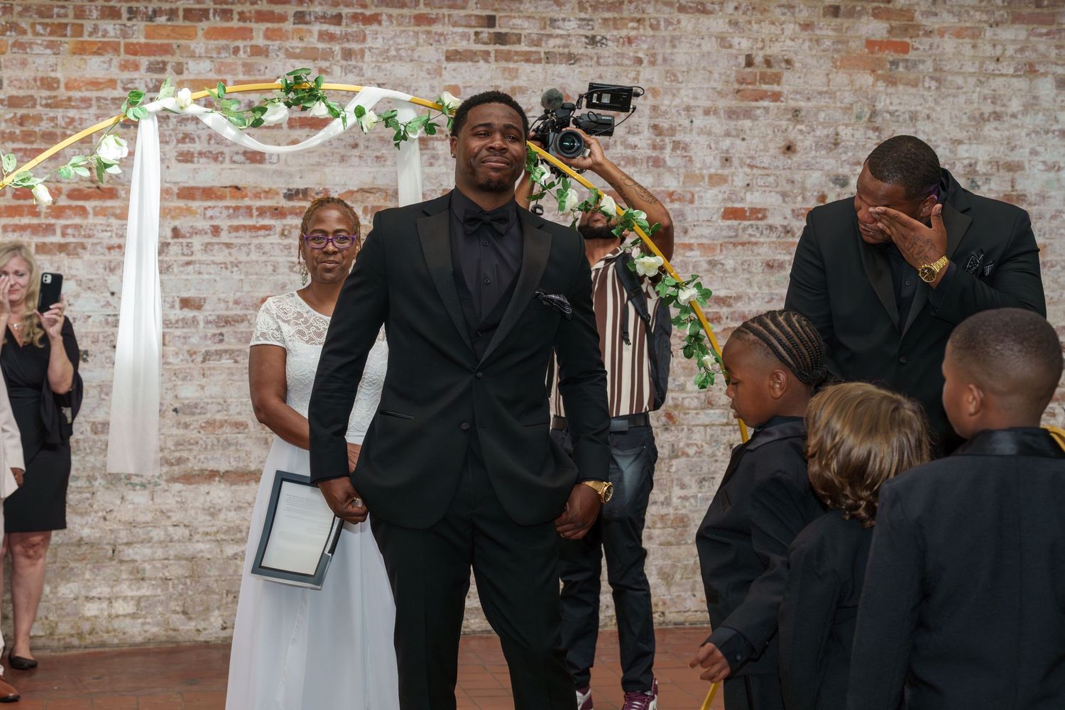 Emotional groom in black tuxedo reacts at wedding altar with guests and officiant in brick venue.